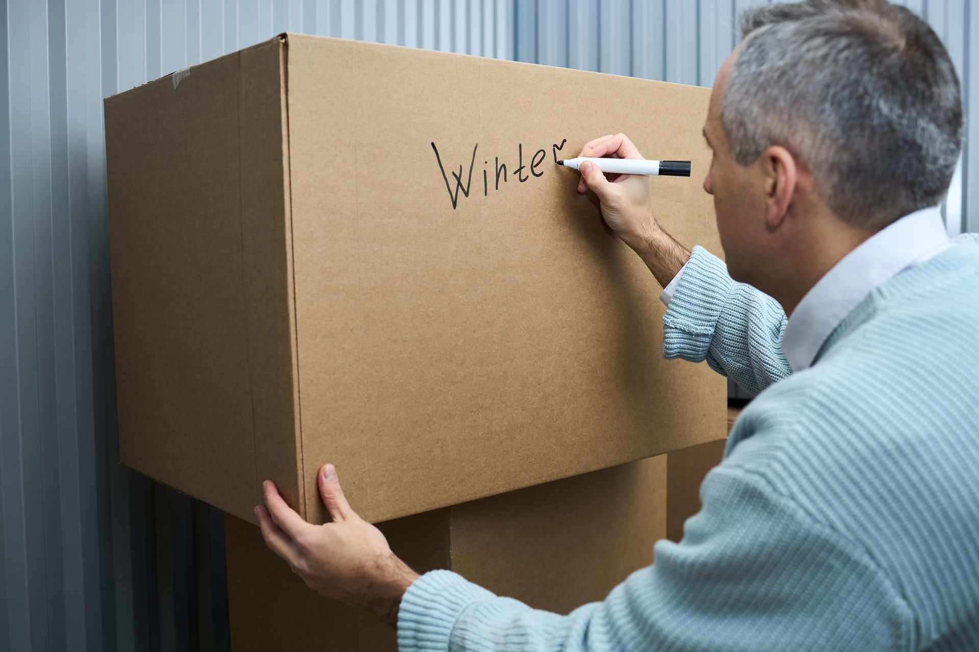 A man is labeling a cardboard box, writing the word 'Winter' on the surface for storage.