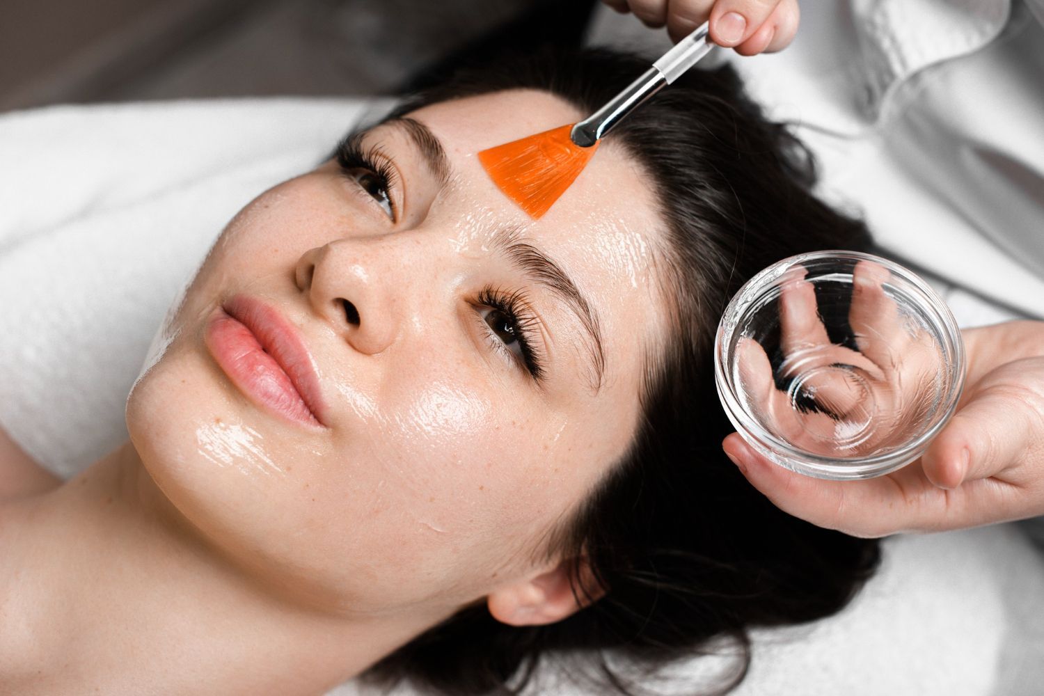 Woman receiving a facial treatment with orange mask applied to forehead and a glass bowl nearby