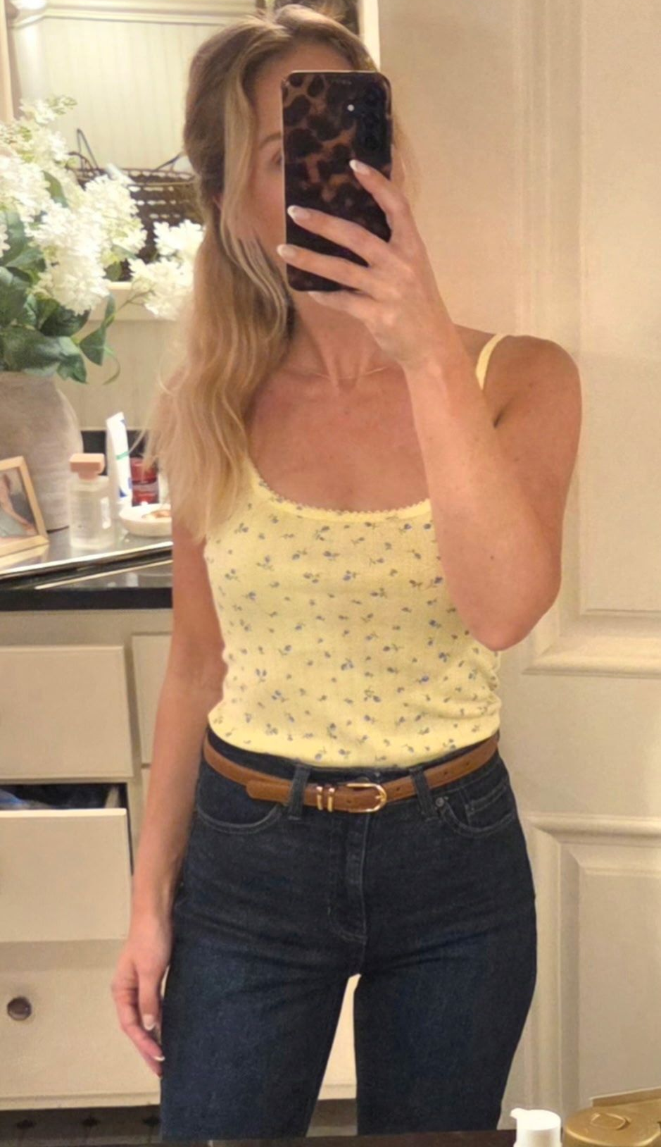 Mirror selfie in a white tank top and dark jeans, holding a phone in a warmly lit room.