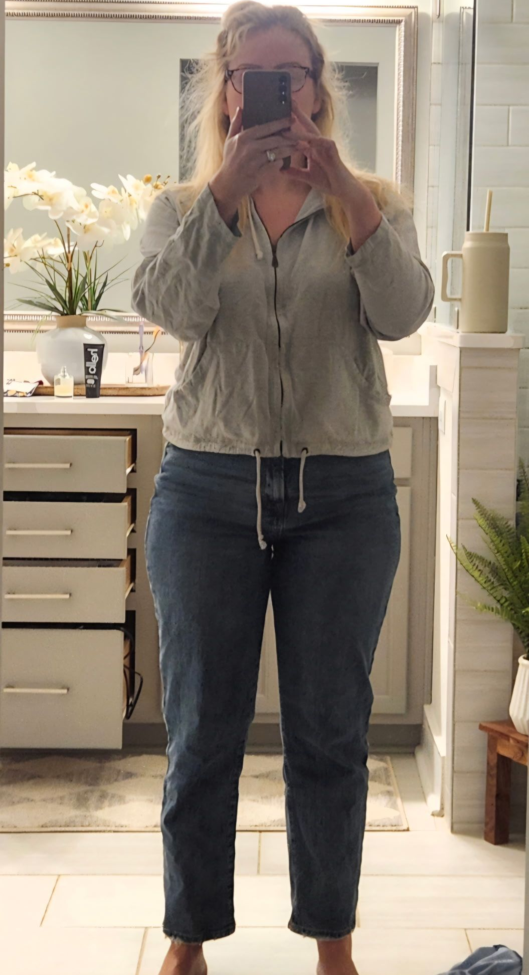 Person taking a mirror selfie in a bright kitchen, wearing a light blouse and jeans.