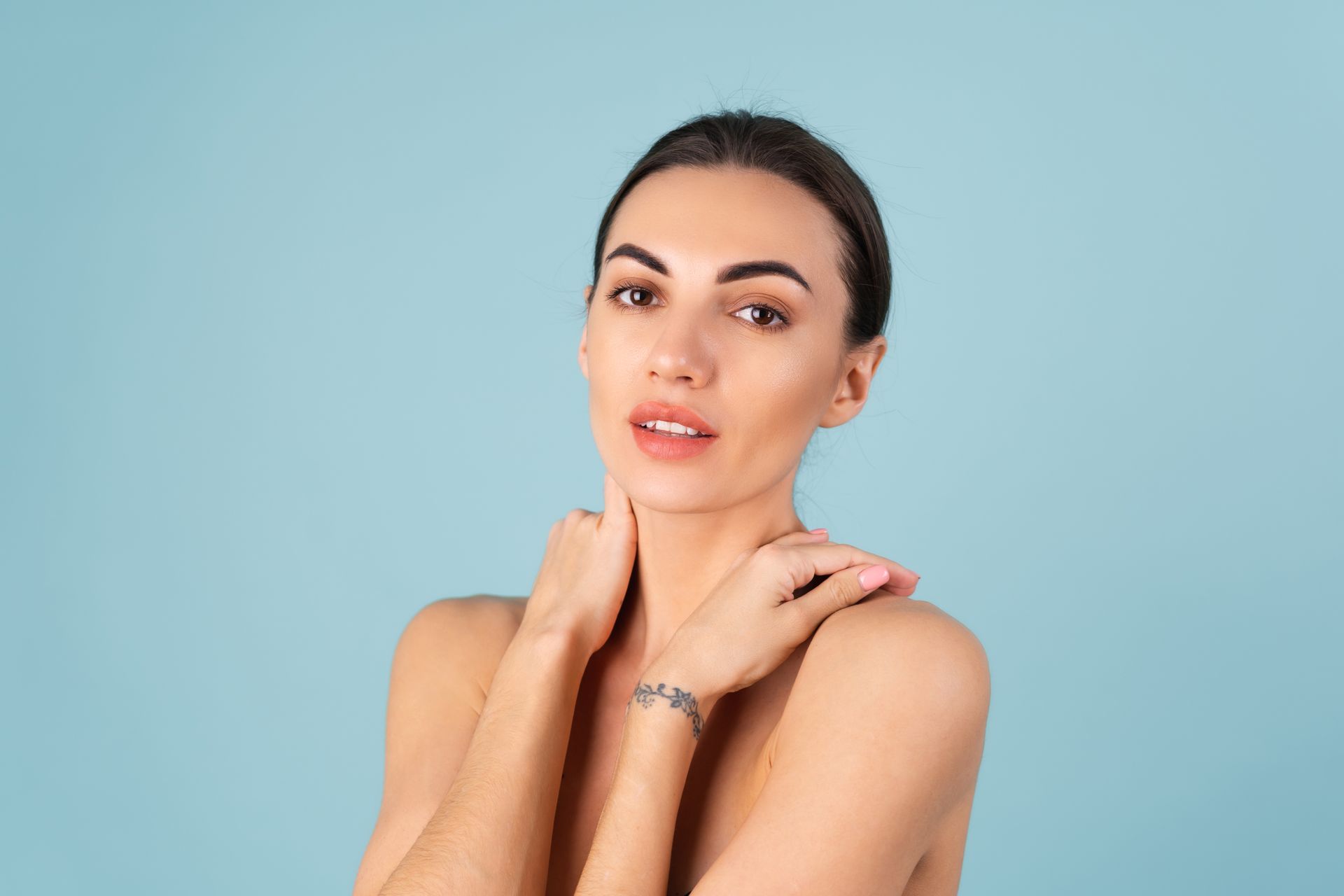 Portrait of a woman against a light blue background, touching her neck and looking at the camera.