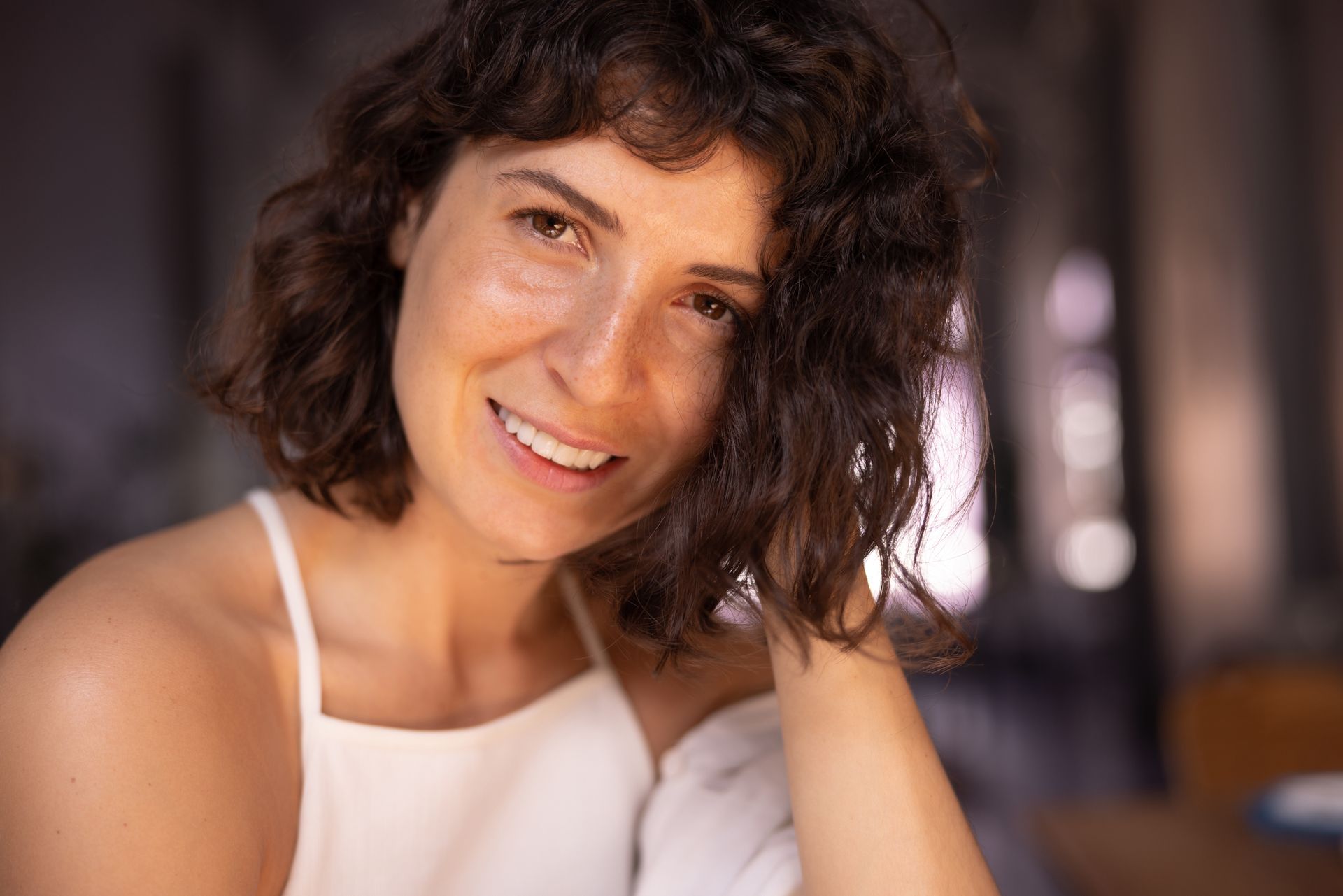 Smiling woman with curly dark hair in a white top, resting her head on her hand indoors.
