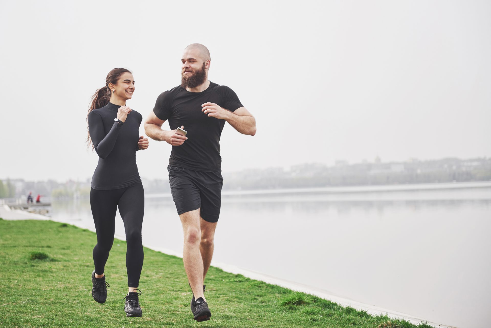 Two people jogging side by side on a grassy path beside a calm lake on a foggy morning