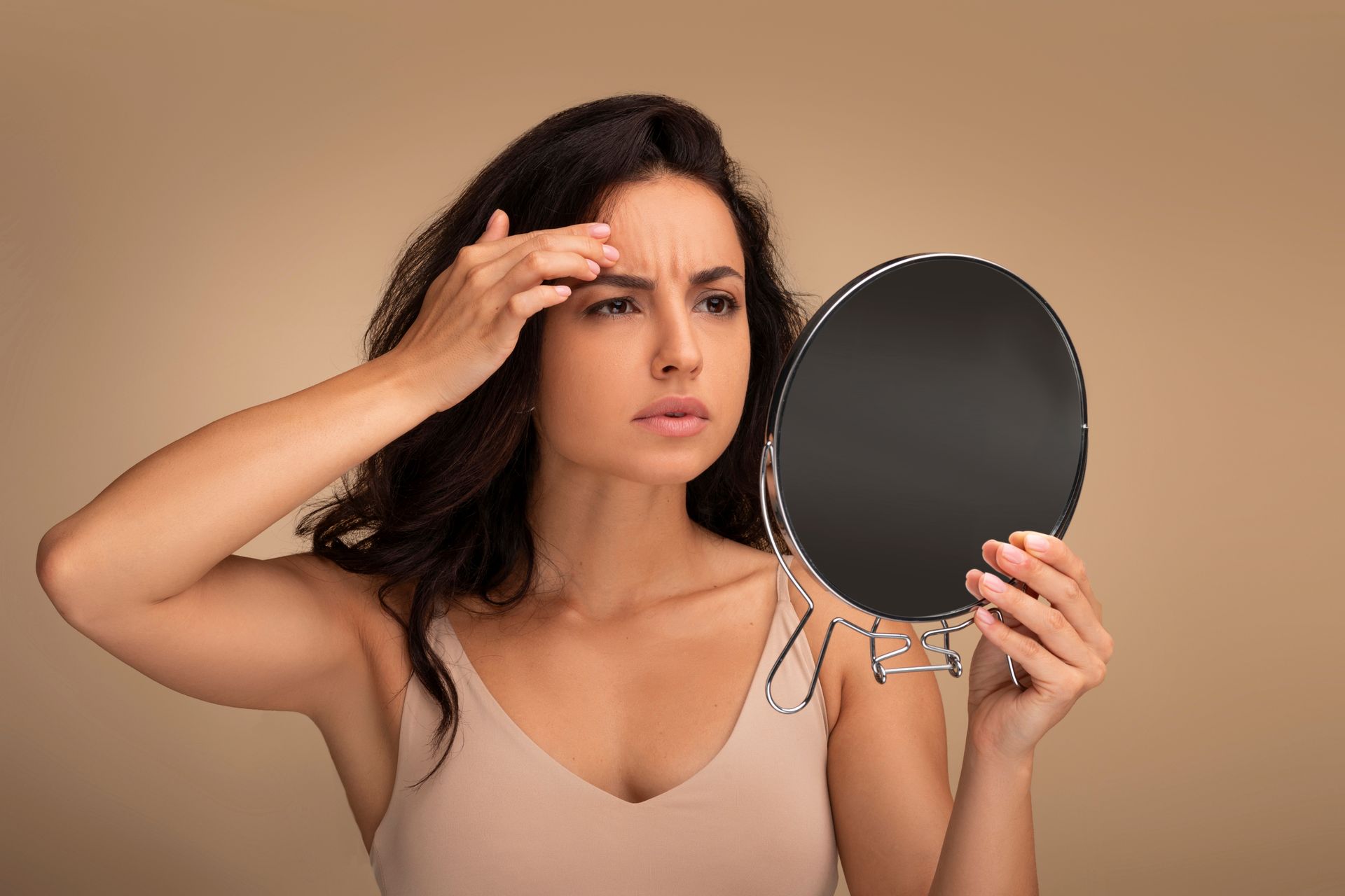 Woman examining her forehead in a mirror, checking for skin concerns on a beige background