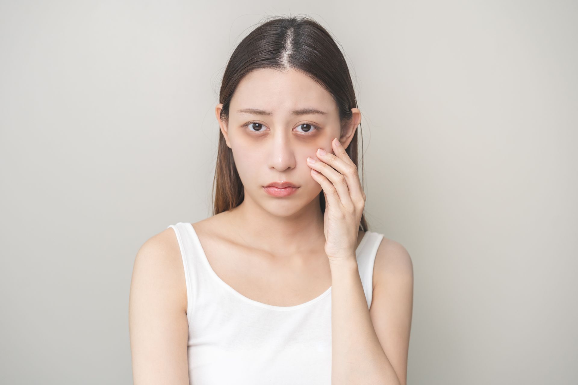 Woman in a white top touching her cheek, looking concerned against a plain background