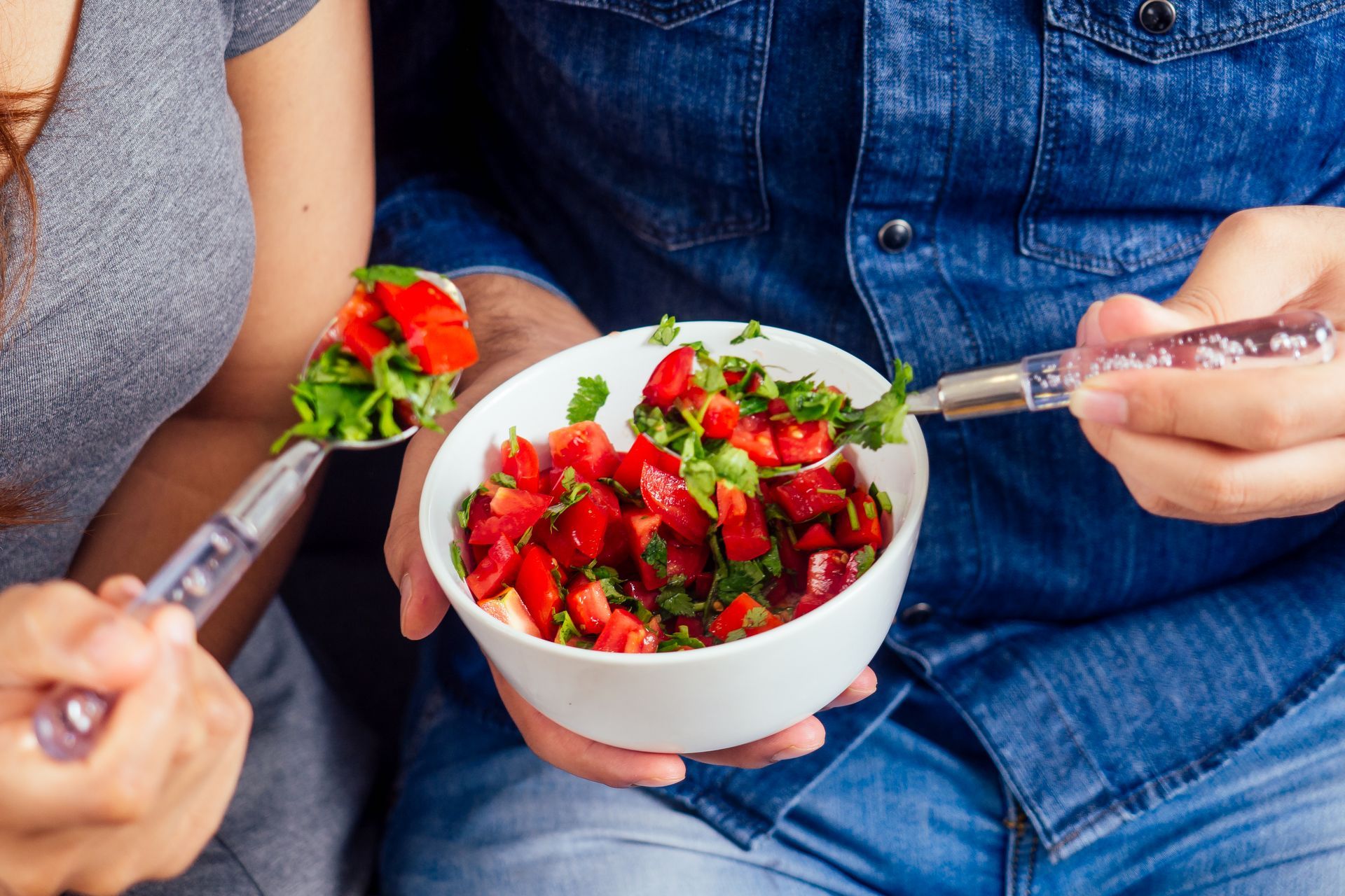 A man and a woman are eating a salad from a bowl with spoons.
