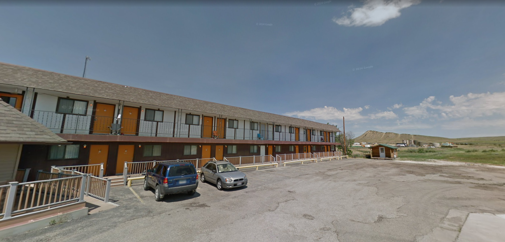 Row of tan townhouses with parked cars on a gravel lot under a cloudy sky