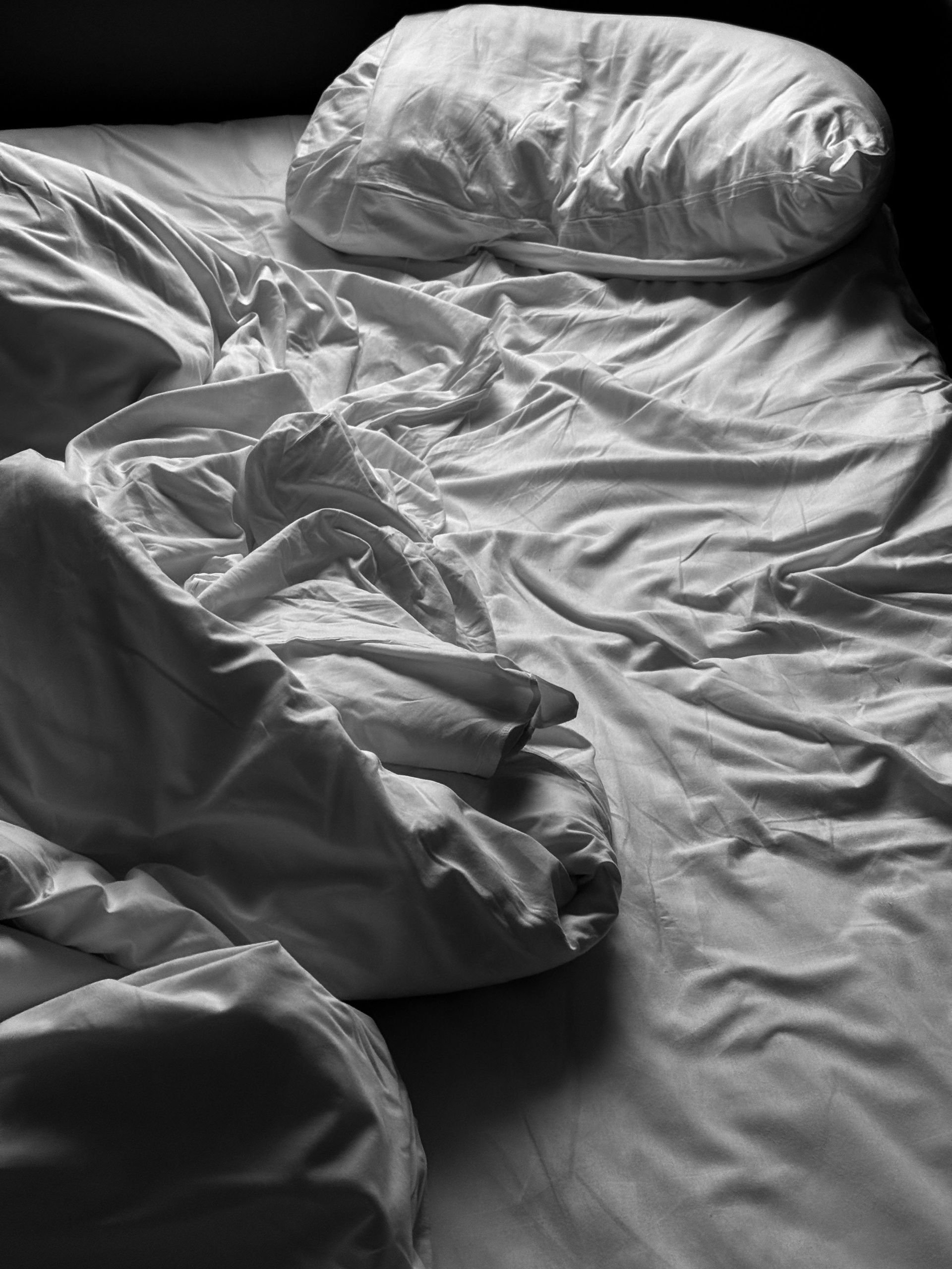 Unmade bed with wrinkled sheets and a pillow in grayscale lighting