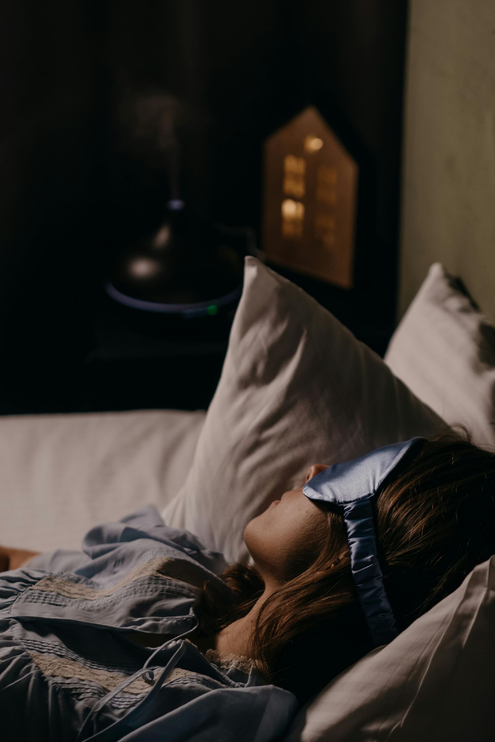 Person sleeping in bed with gray pillows and blanket, lit by a warm lamp in a dark room