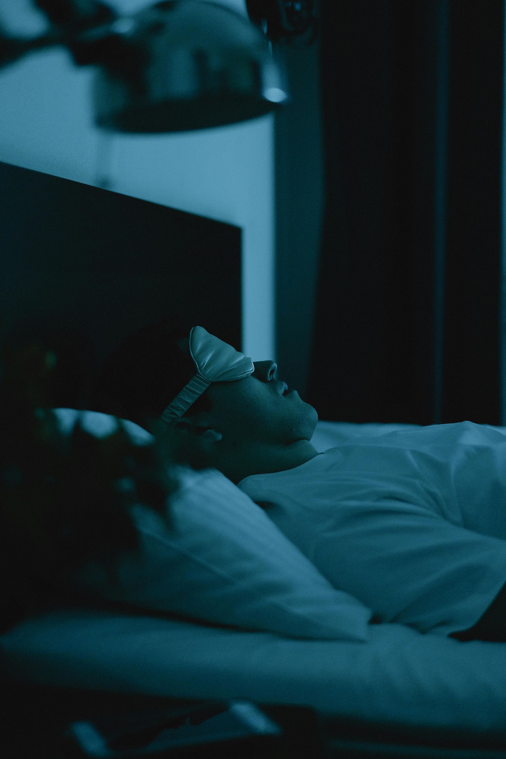 Person lying in bed at night, lit by blue light, with a hand on their forehead.