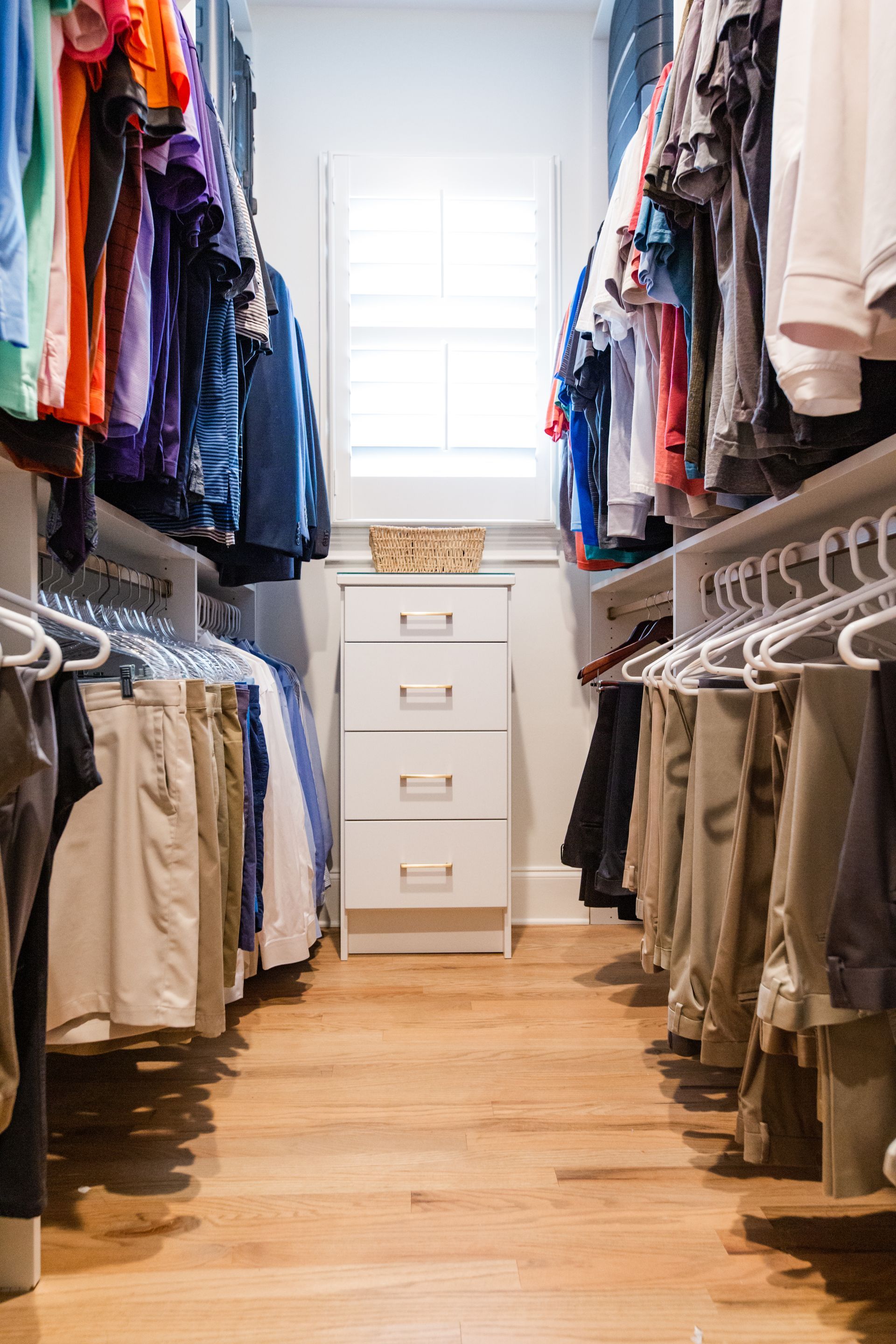 Walk-in closet with clothes hanging on both sides, a white dresser, and a window. Wooden floors.