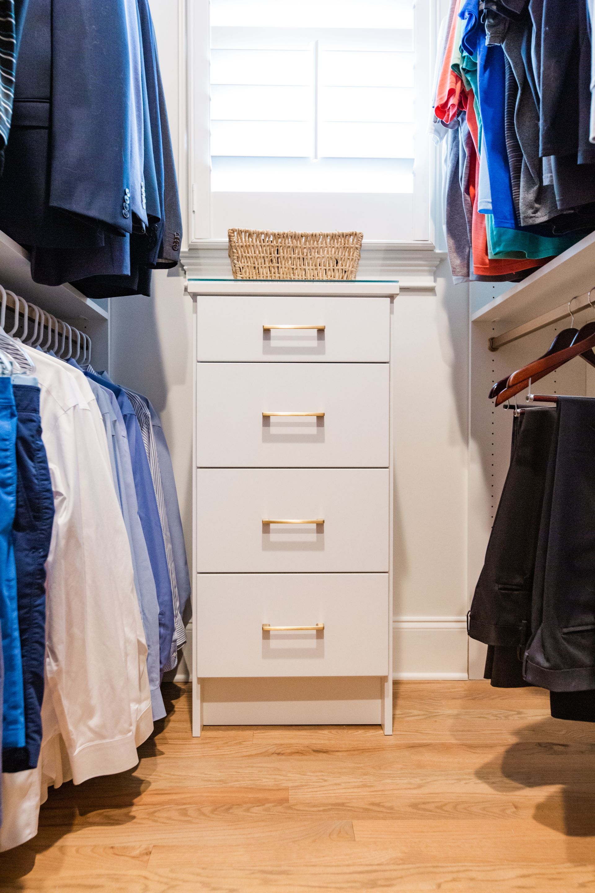 Walk in Closet with white drawers and hanging clothes, wood floor. Light streams in from window above drawers.