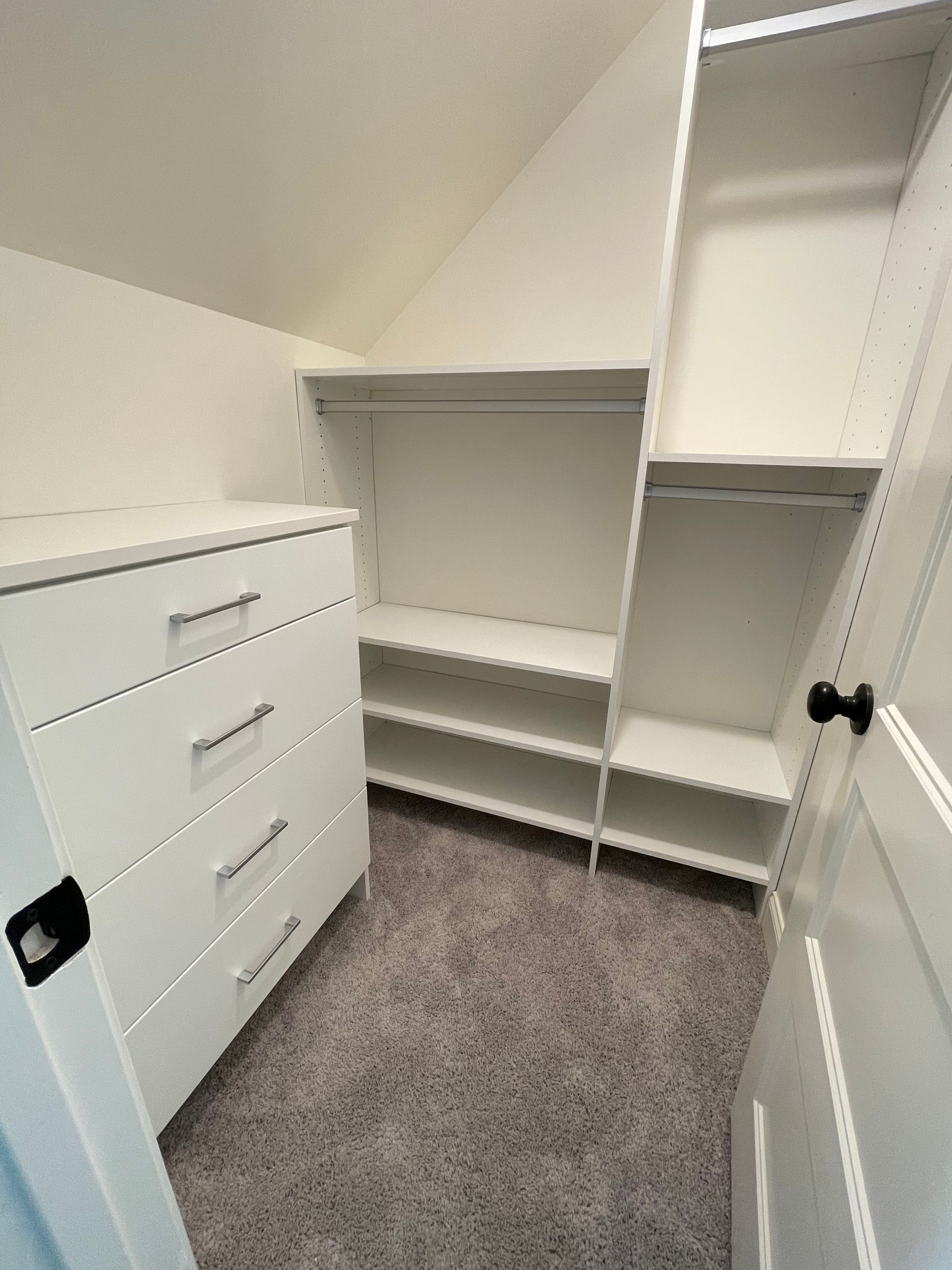 White closet with shelves, drawers, and a clothing rod, on gray carpet. Angled ceiling.