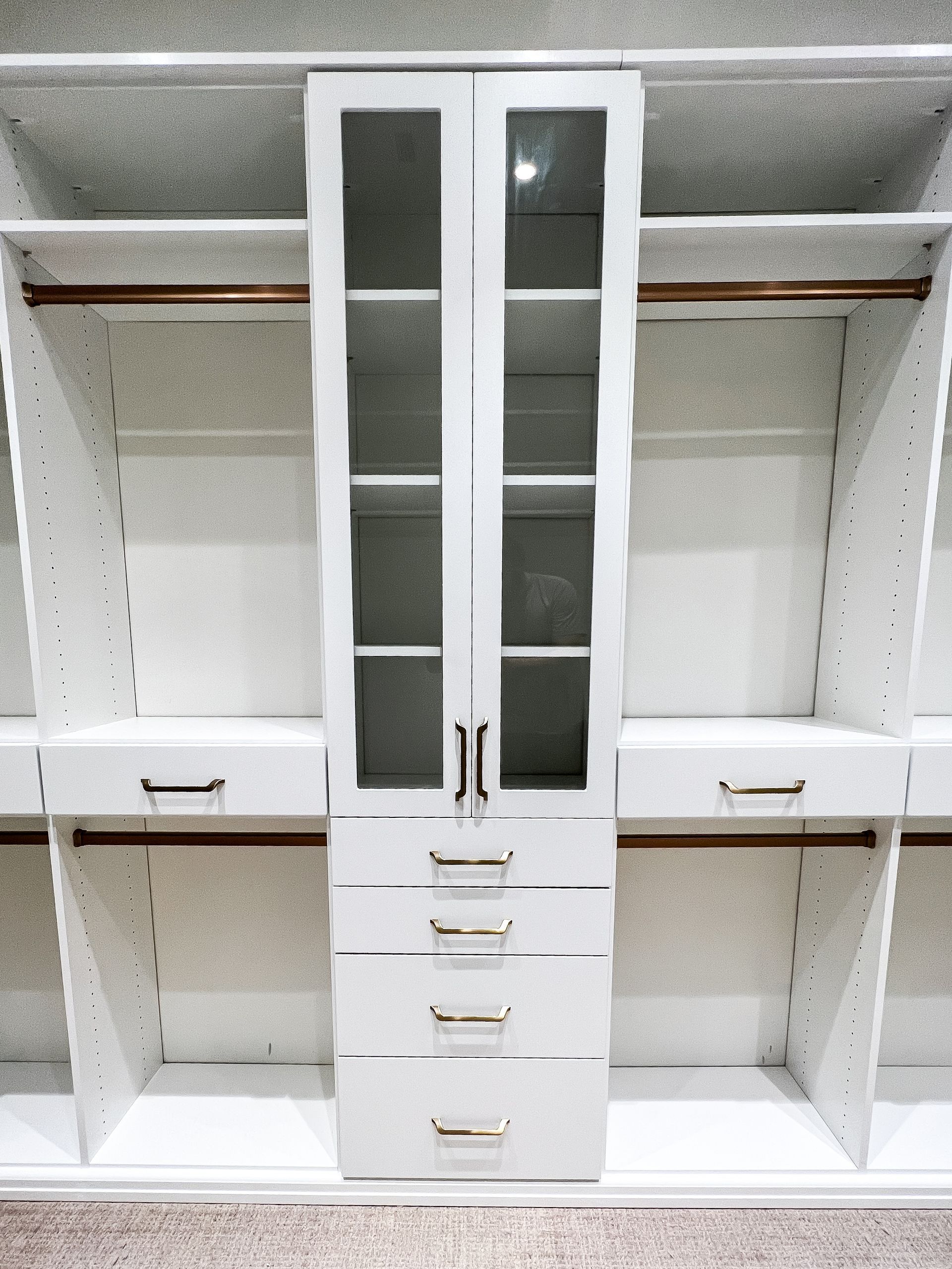 White, custom closet with center glass-door cabinet, drawers, and surrounding shelves with hanging rods.
