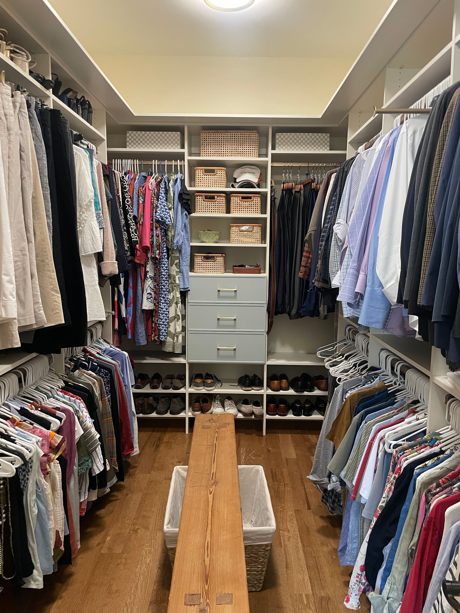 Walk-in closet with clothes on racks, shelves with boxes, and shoes below. Wooden bench in the middle.