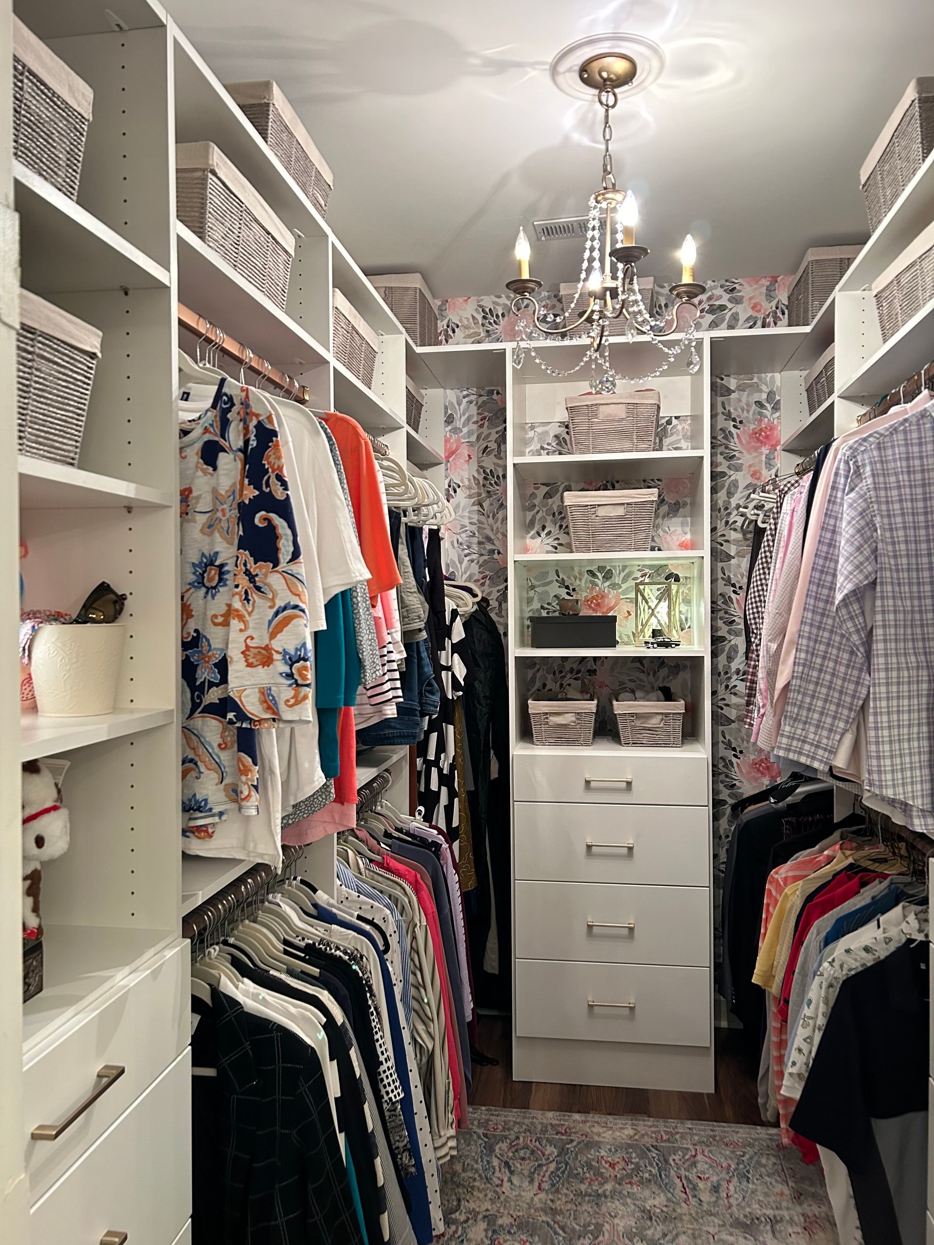 Walk-in closet with white shelves, hanging clothes, decorative wallpaper, and chandelier.