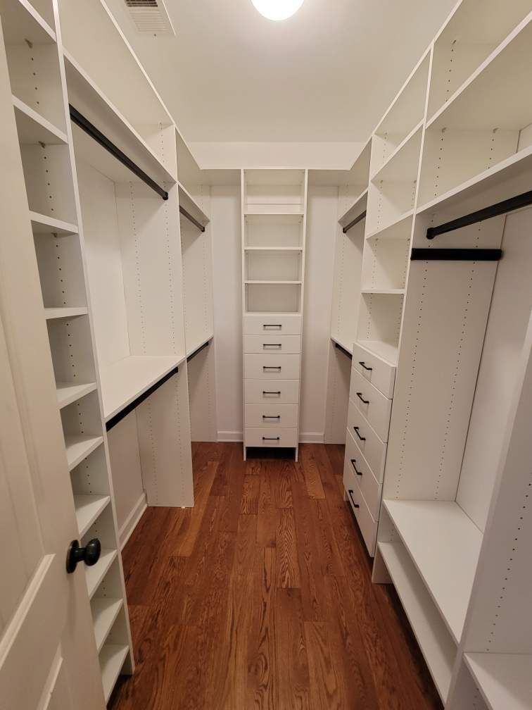 Walk-in closet with white shelving, black rods, drawers, and wooden floor.