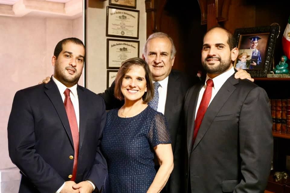 Four people pose indoors, dressed formally, with certificates and framed photos in the background.
