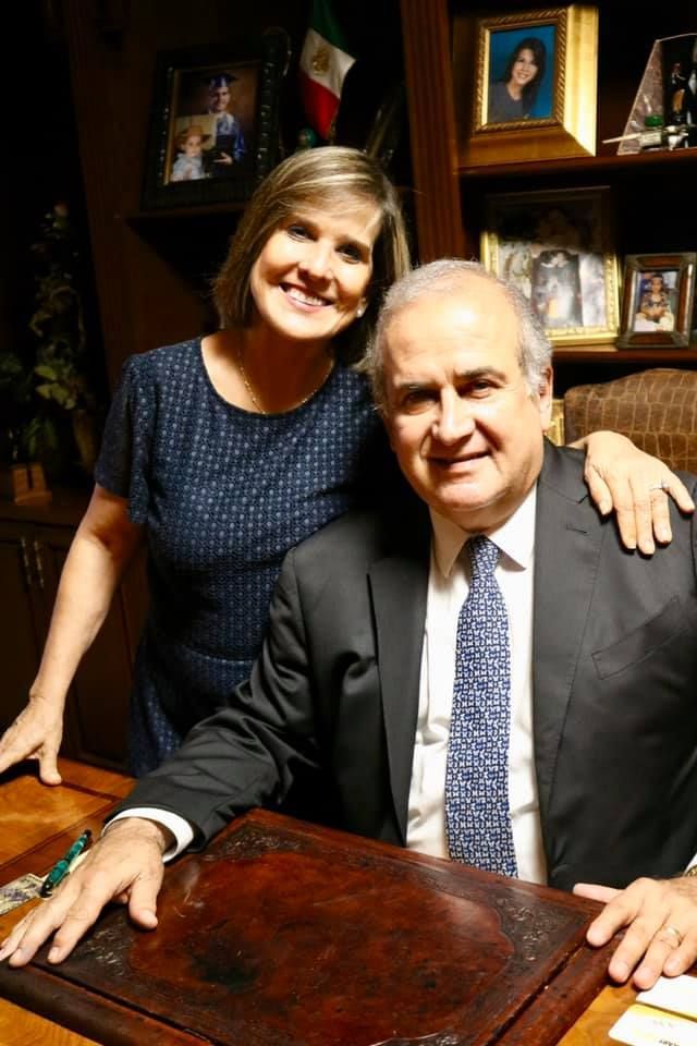 Two people smiling behind a wooden desk in a framed-photo office setting.