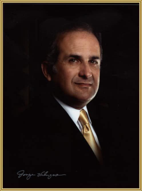 Formal portrait of a man in a dark suit and gold tie against a black background.