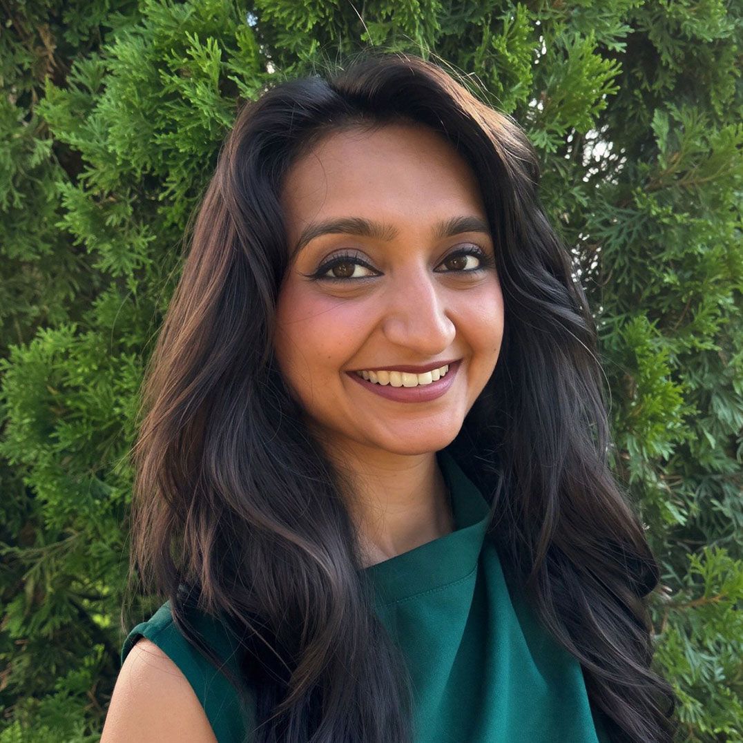 Smiling person with long dark hair standing outdoors before green trees