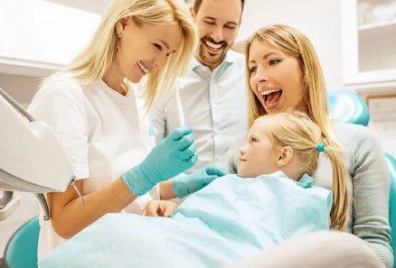 Dentist and family smiling beside a child in a dental chair, with a turquoise bib and tools nearby
