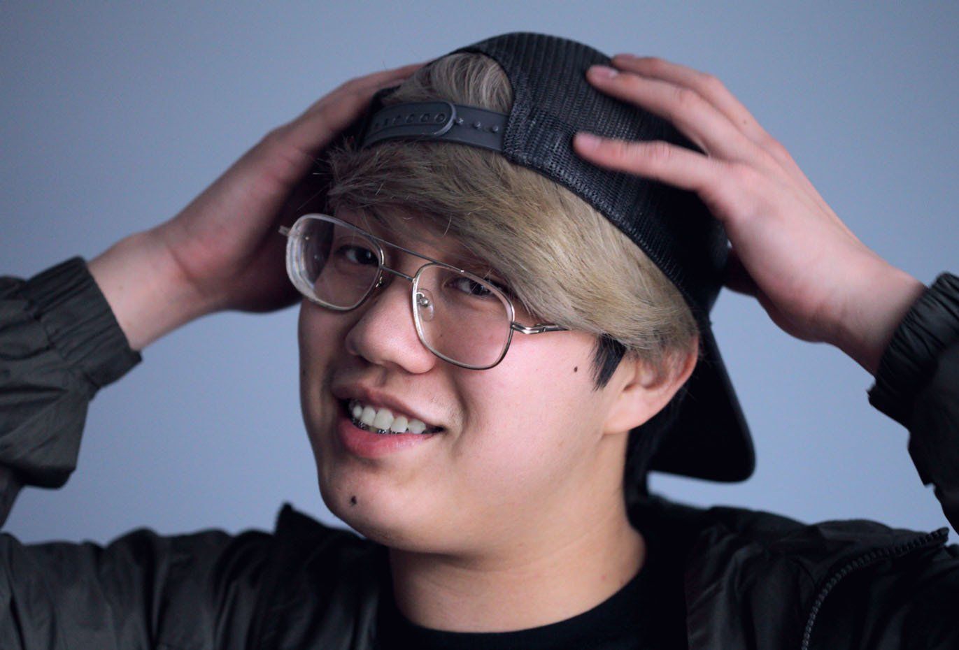Person with glasses and blond hair, smiling while adjusting a black cap against a gray background