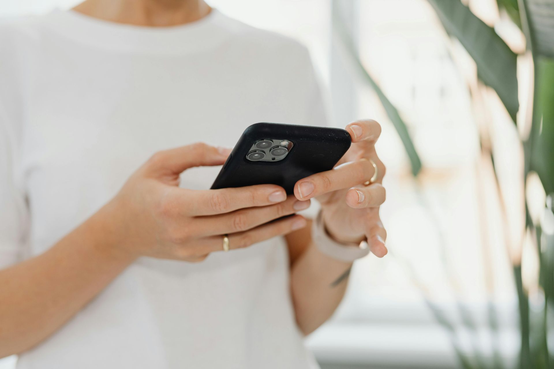 Woman holding a black smartphone with both hands, wearing a white shirt and rings, near a green plant.