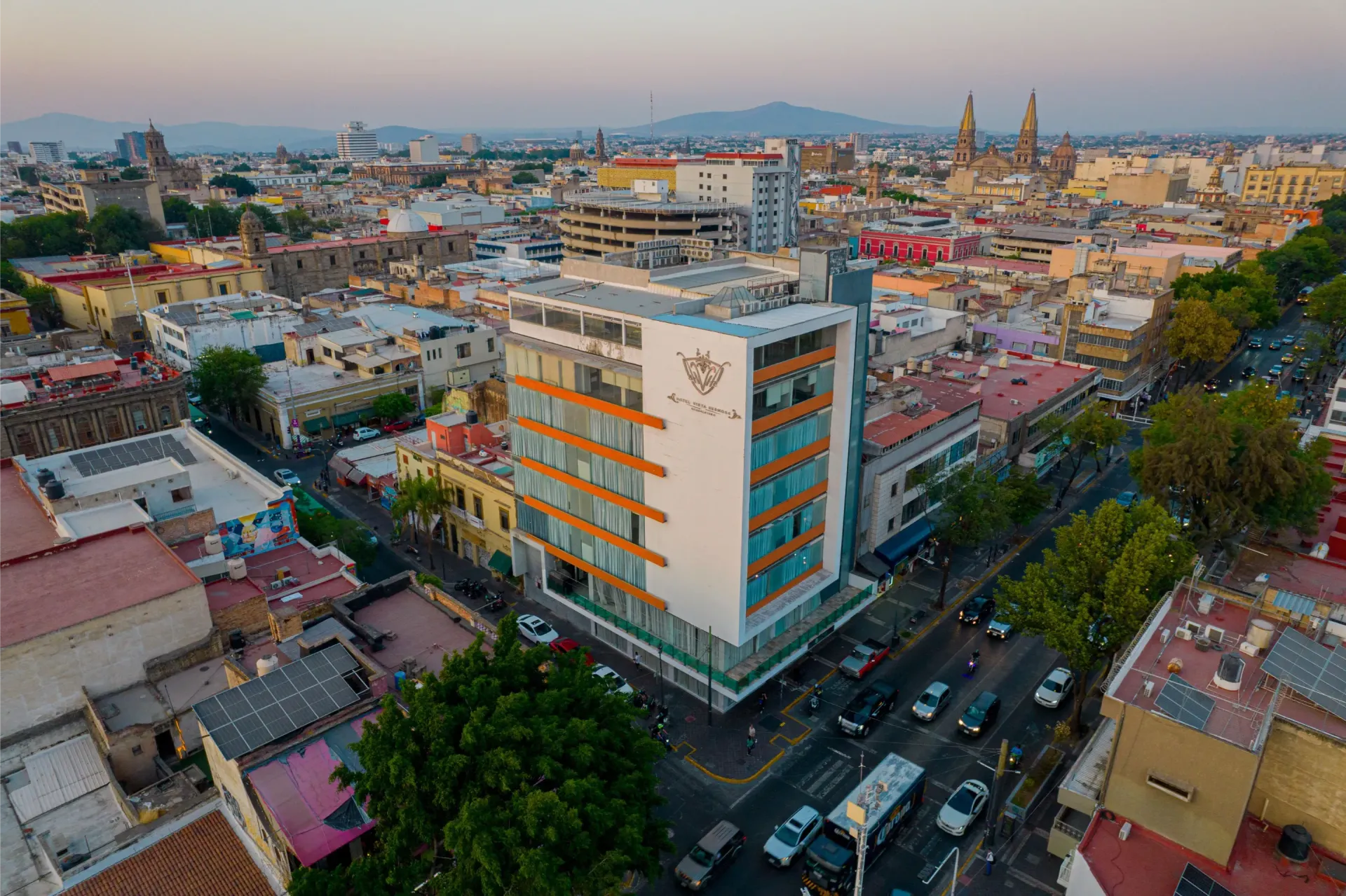 Vista aérea de una manzana con un edificio blanco y naranja, calles concurridas y, a lo lejos, una catedral en el horizonte.