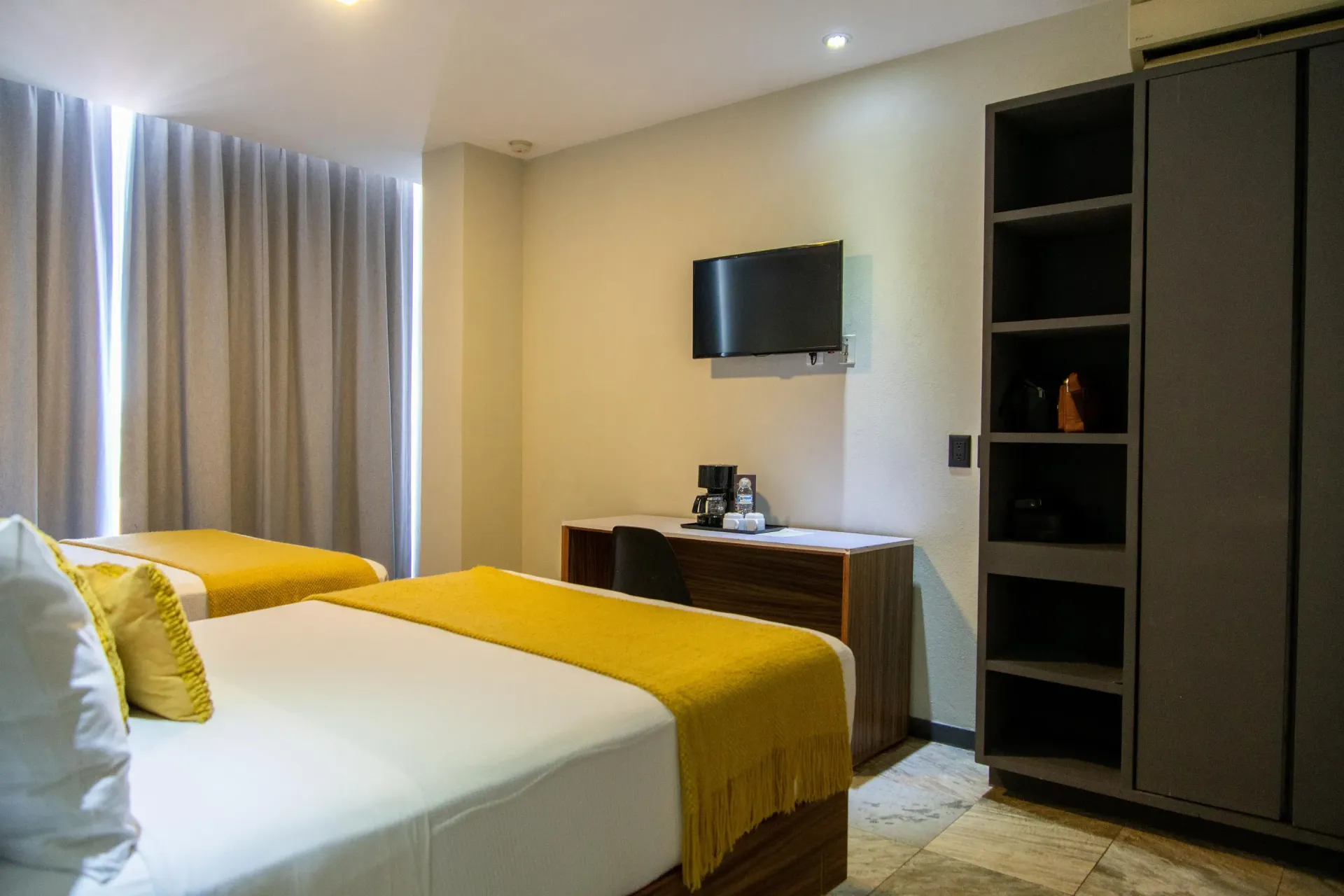 Dormitorio moderno con dos camas, televisor montado en la pared, cómoda y armario gris, con detalles en amarillo y blanco.