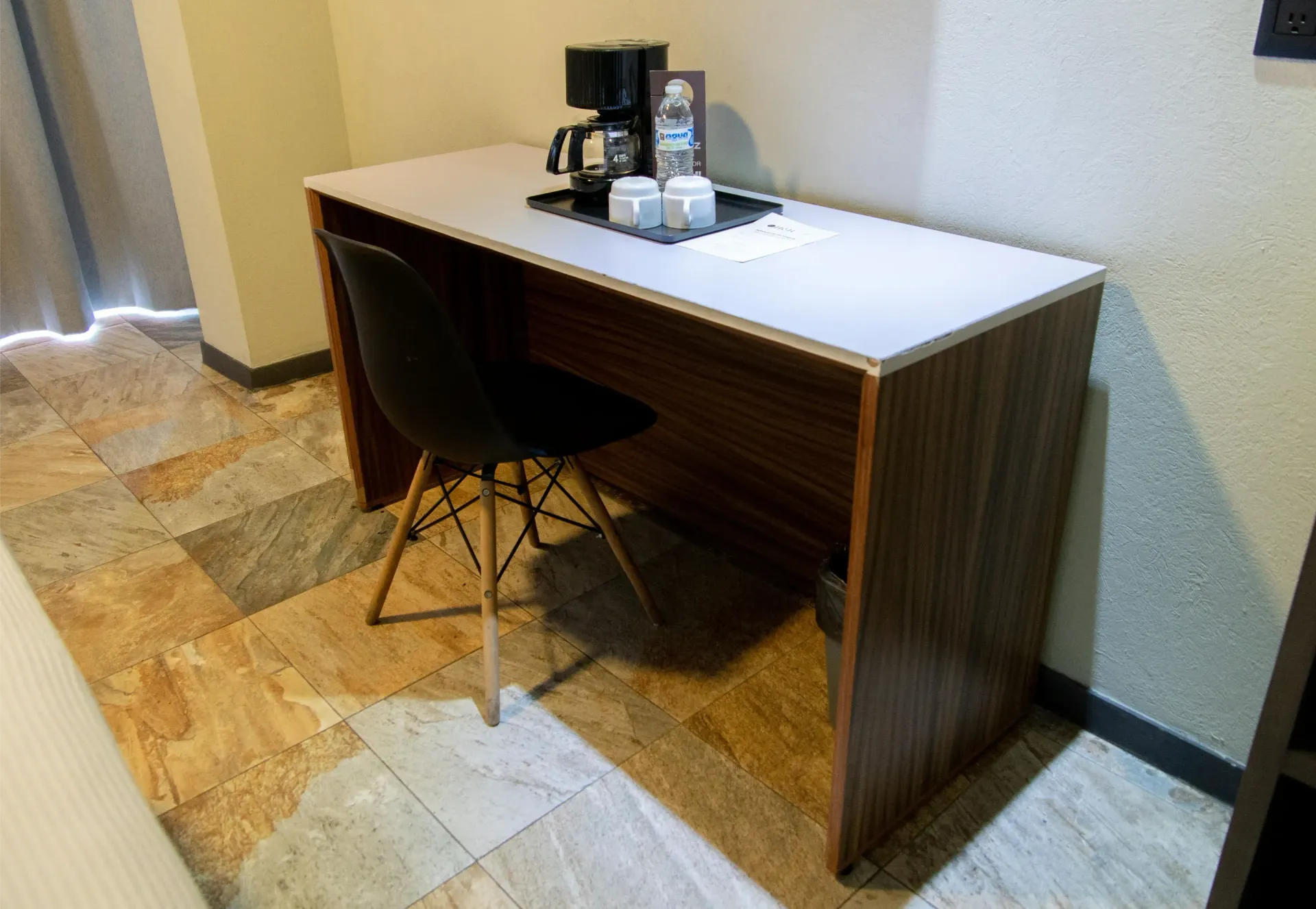 Pequeño mostrador de hotel con una silla negra, cafetera y tazas en una habitación con suelo de baldosas.