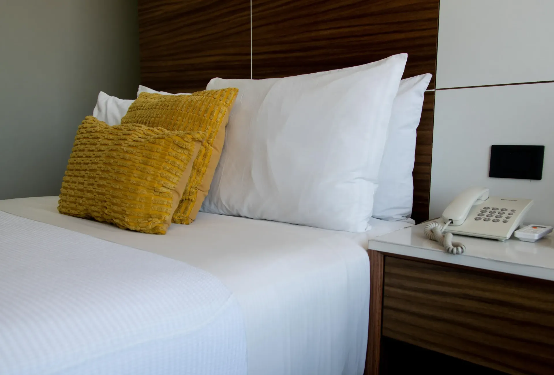 Cama de hotel con sábanas blancas, cojines decorativos amarillos y un teléfono de cabecera sobre una mesita de noche.