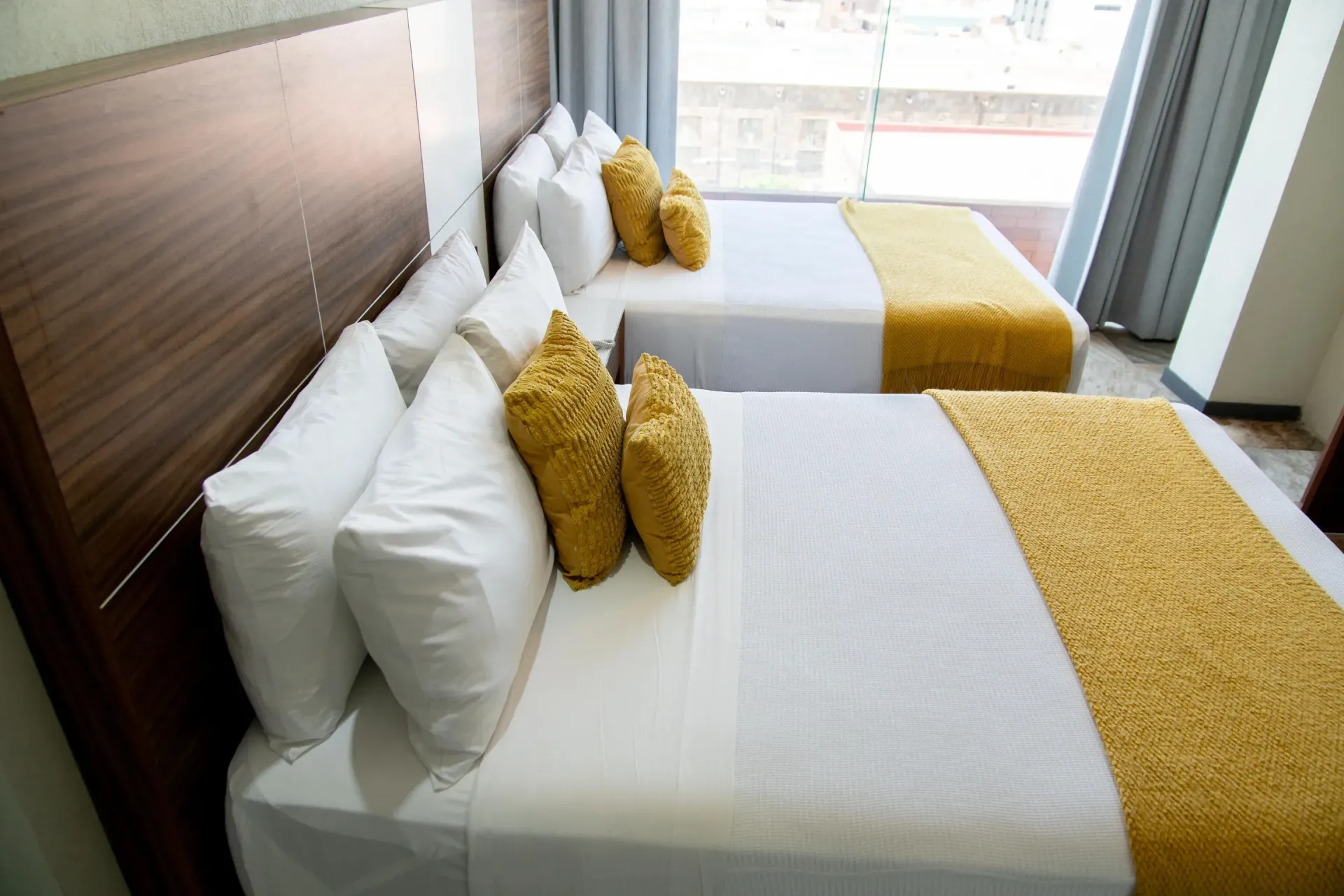 Tres camas bien hechas con sábanas blancas y almohadas amarillas en una luminosa habitación de hotel.