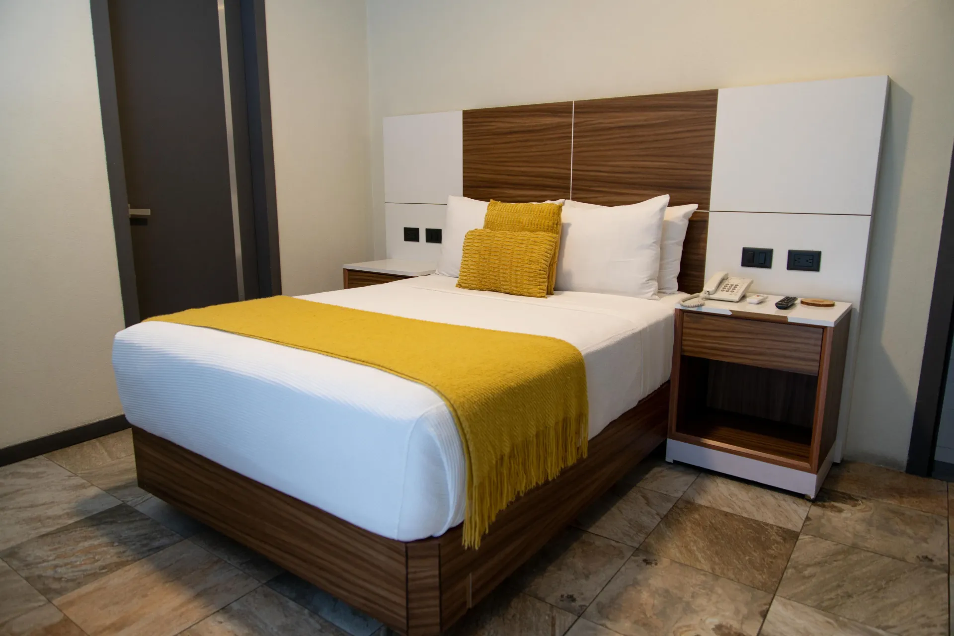 Habitación de hotel moderna con una cama bien hecha, un camino de mesa amarillo y mesitas de noche a juego.