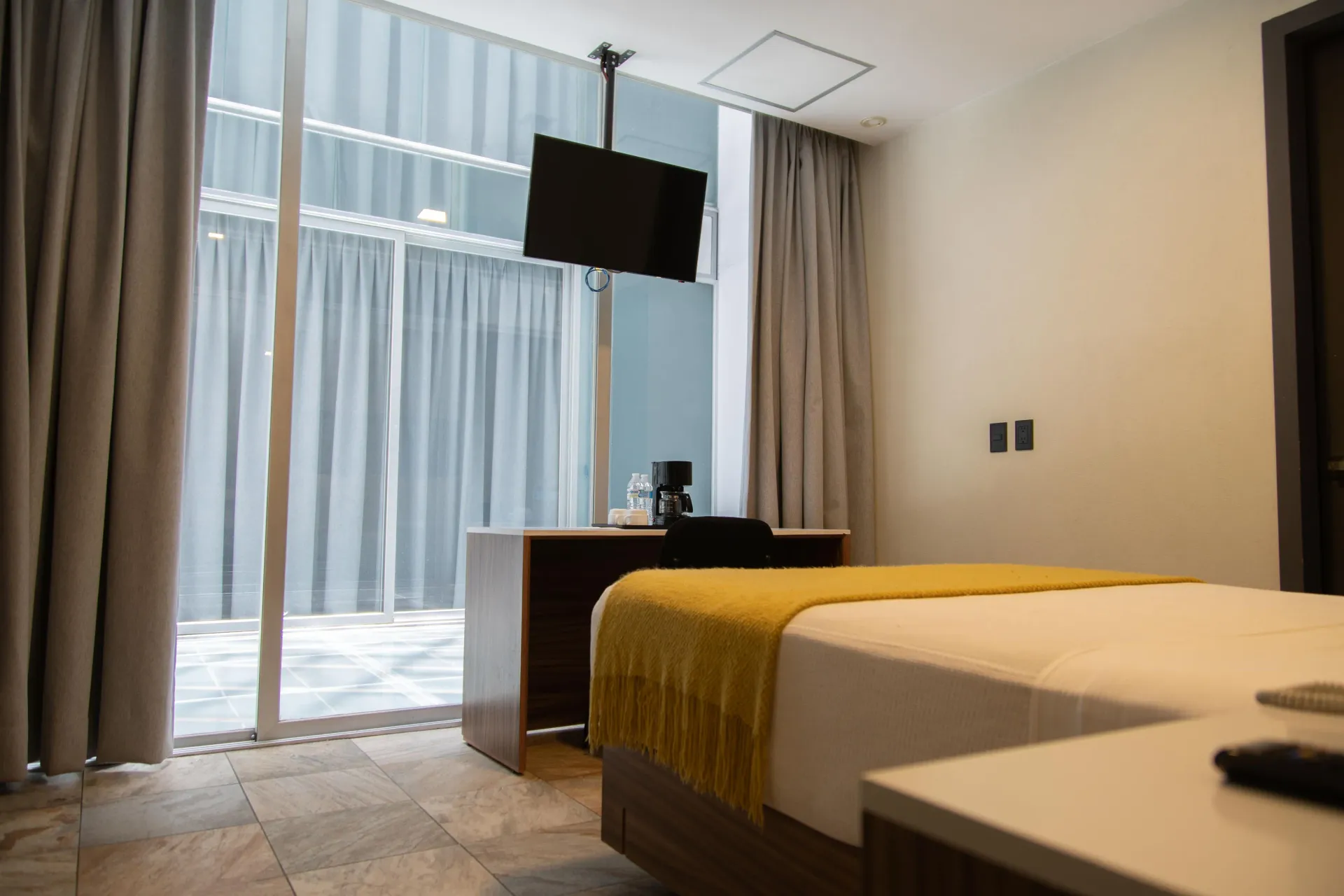 Habitación de hotel moderna con cama, escritorio, televisor de pared y grandes ventanales del suelo al techo con cortinas.