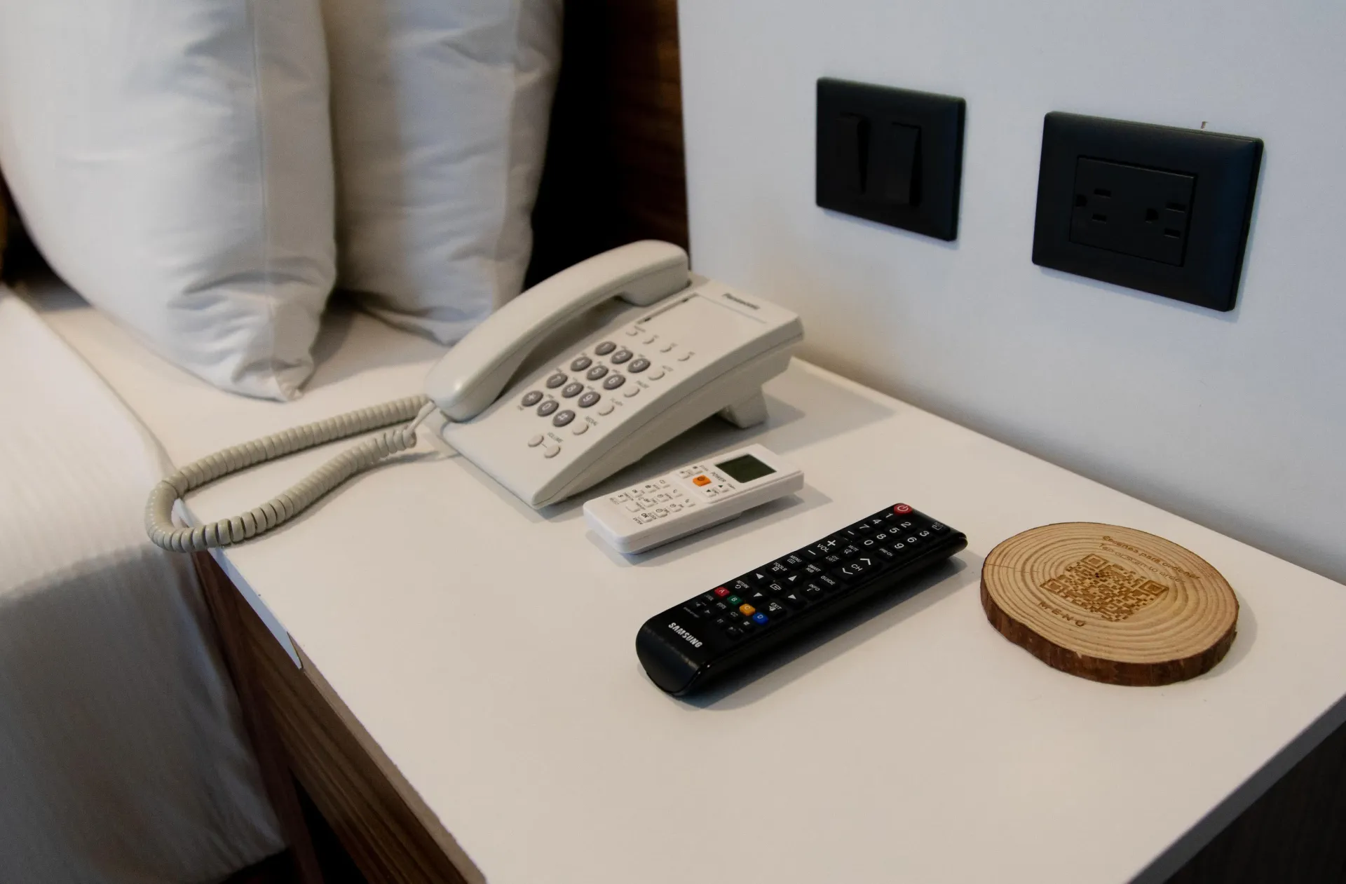 Mesita de noche de hotel con teléfono fijo blanco, mandos a distancia y posavasos junto a la cama y enchufes de pared.