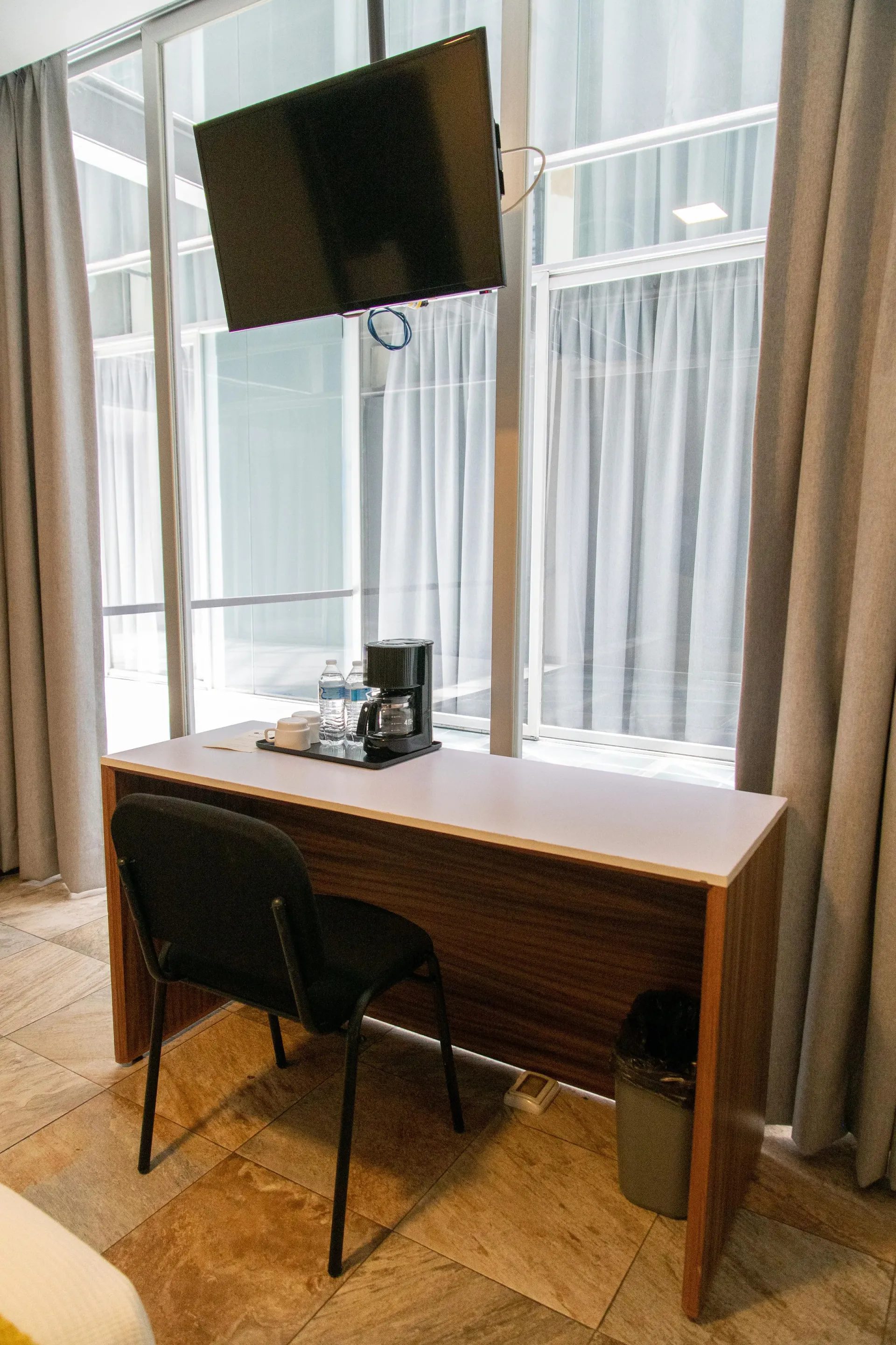 Escritorio pequeño con un televisor montado en la pared frente a ventanas altas y cortinas.