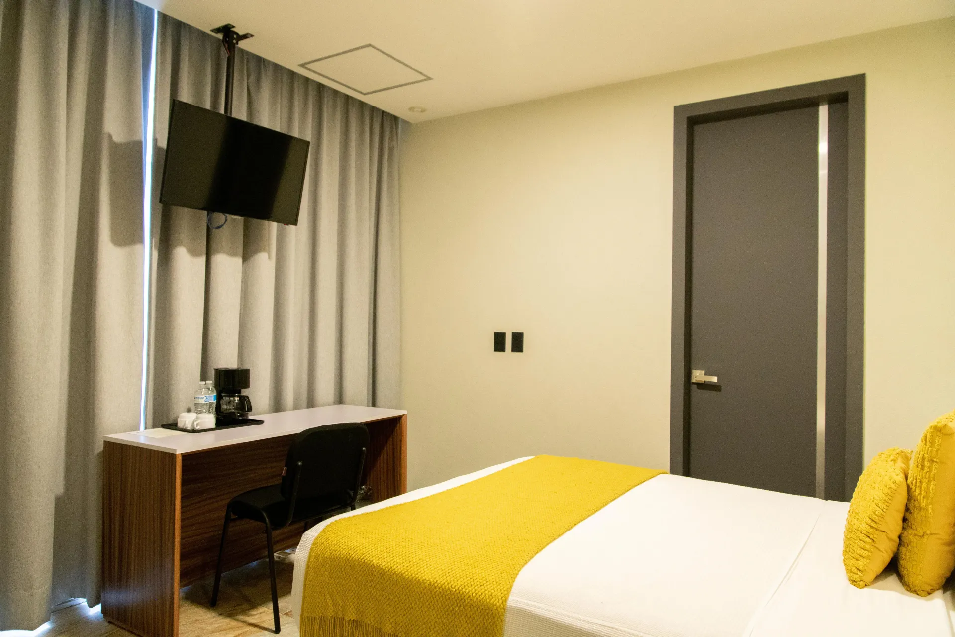 Habitación de hotel pequeña con cama, escritorio, televisor montado en la pared, cortinas y puerta cerrada.