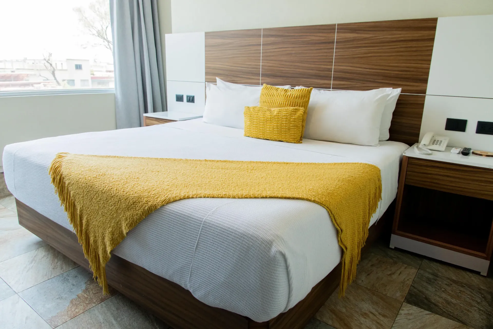 Cama de hotel bien hecha con sábanas blancas, manta y almohada amarillas, junto a mesitas de noche de madera.