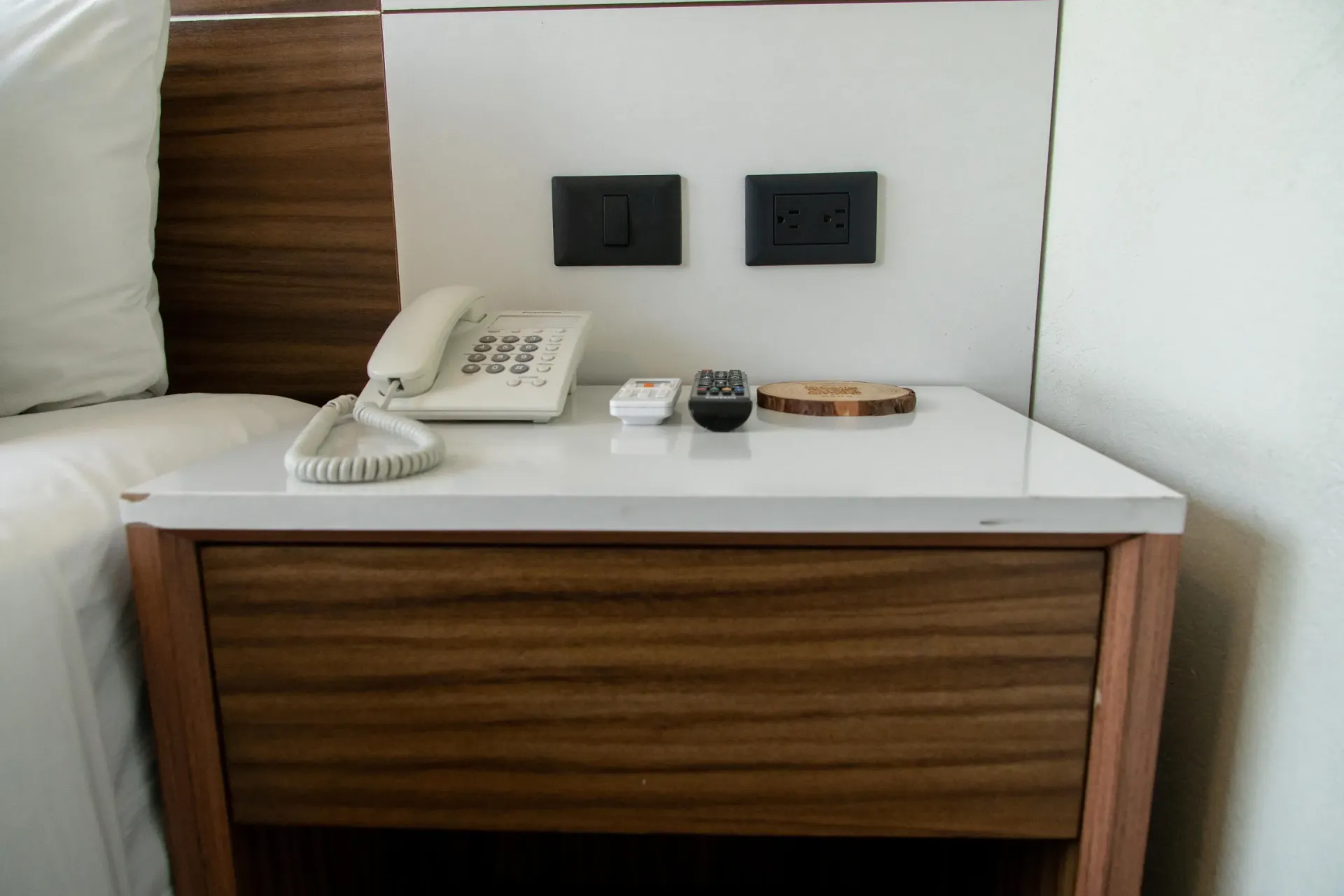 Mesita de noche de hotel con teléfono, lámpara, enchufes y mando a distancia sobre una superficie blanca y paneles de madera.