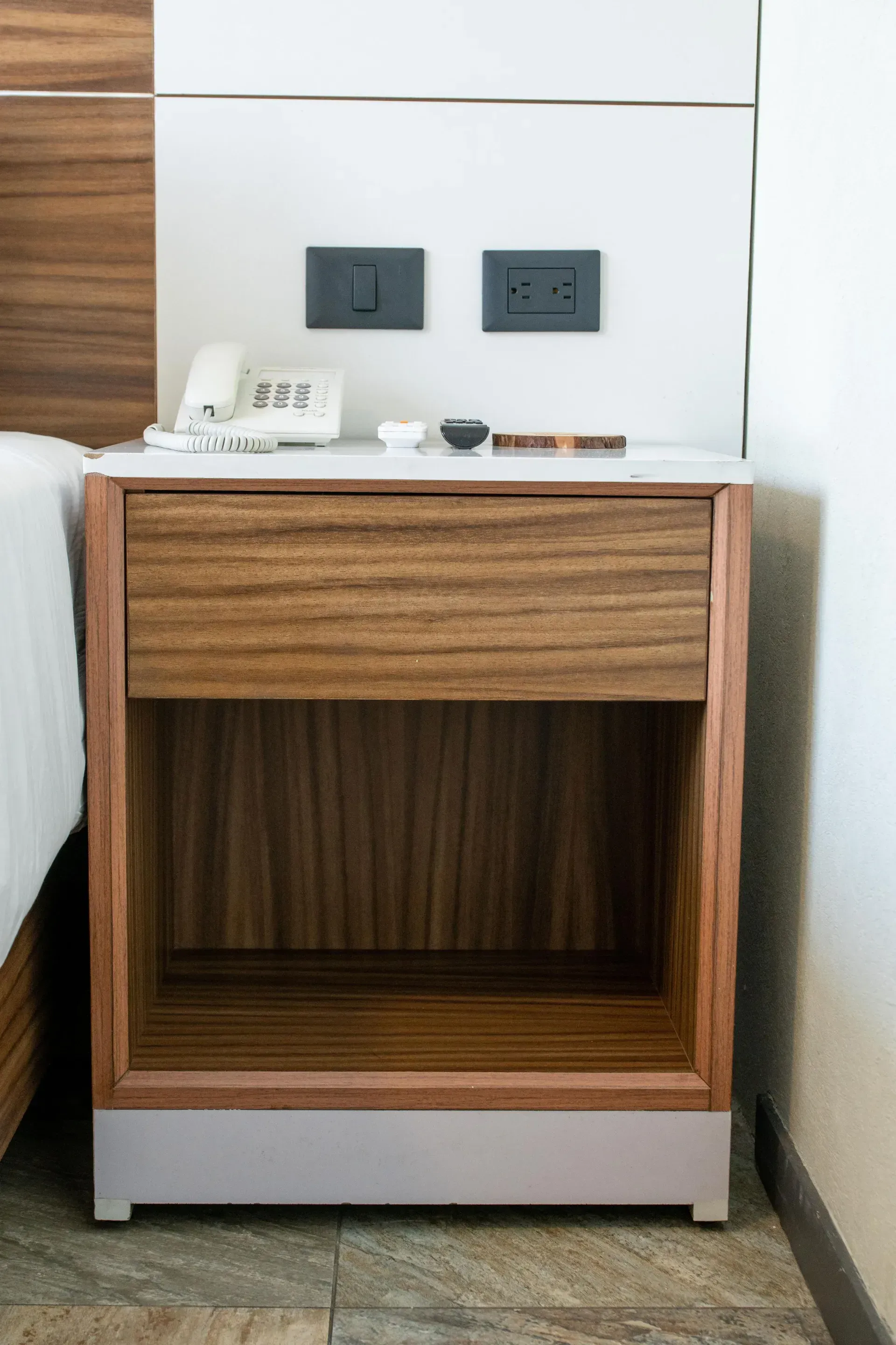 Mesita de noche moderna de madera con cajón, estante, lámpara y enchufes de pared, situada junto a la cama.