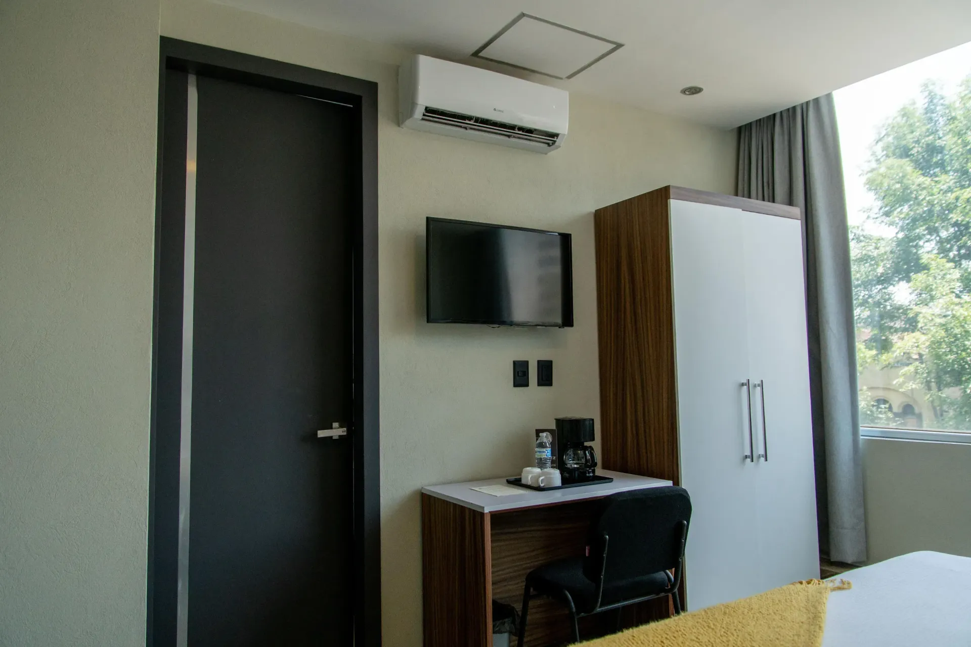 Habitación de hotel con puerta oscura, televisor montado en la pared, escritorio, armario y ventana con cortinas.