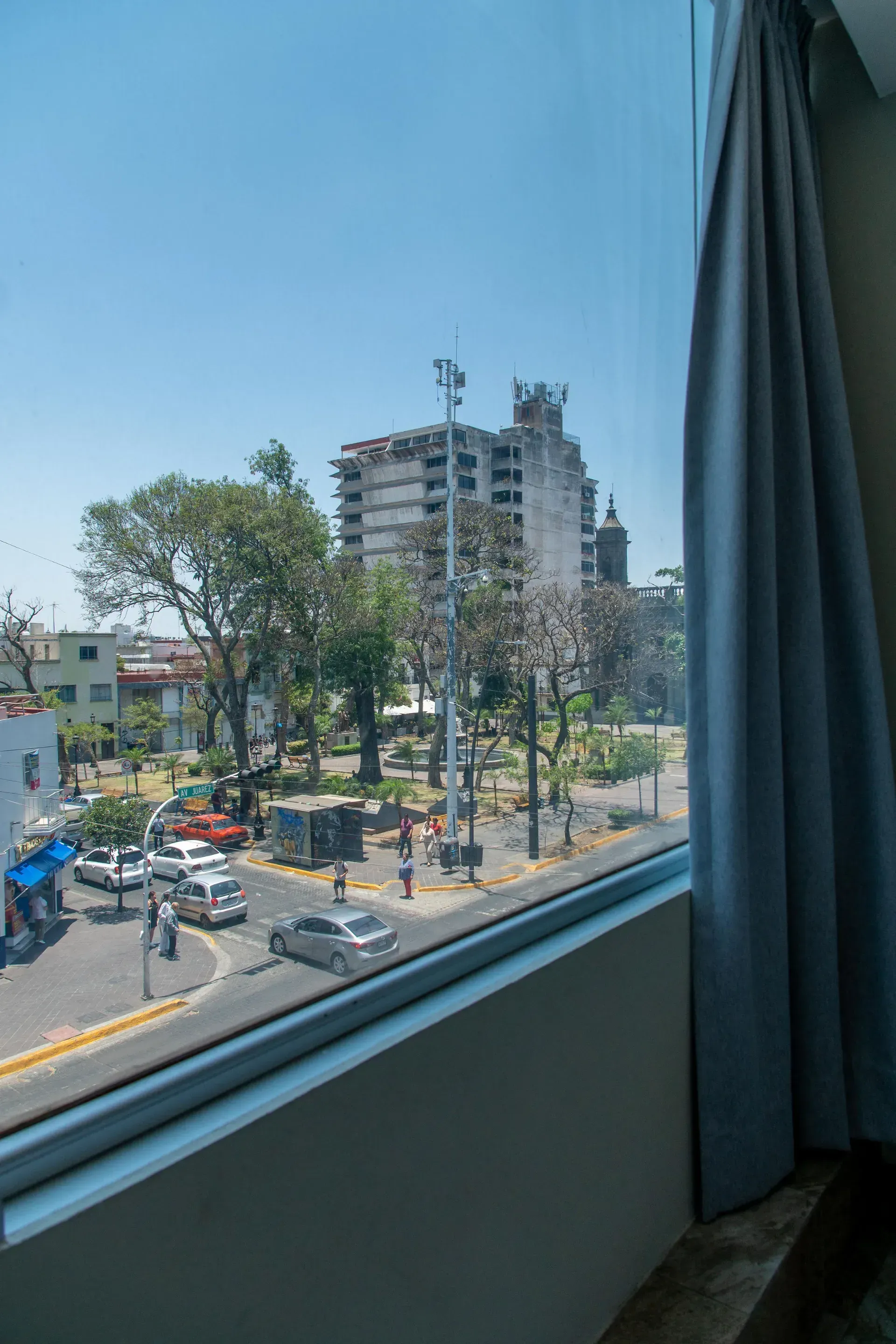 Vista a través de una ventana de una soleada intersección urbana con edificios, árboles y coches; cortina a la derecha.