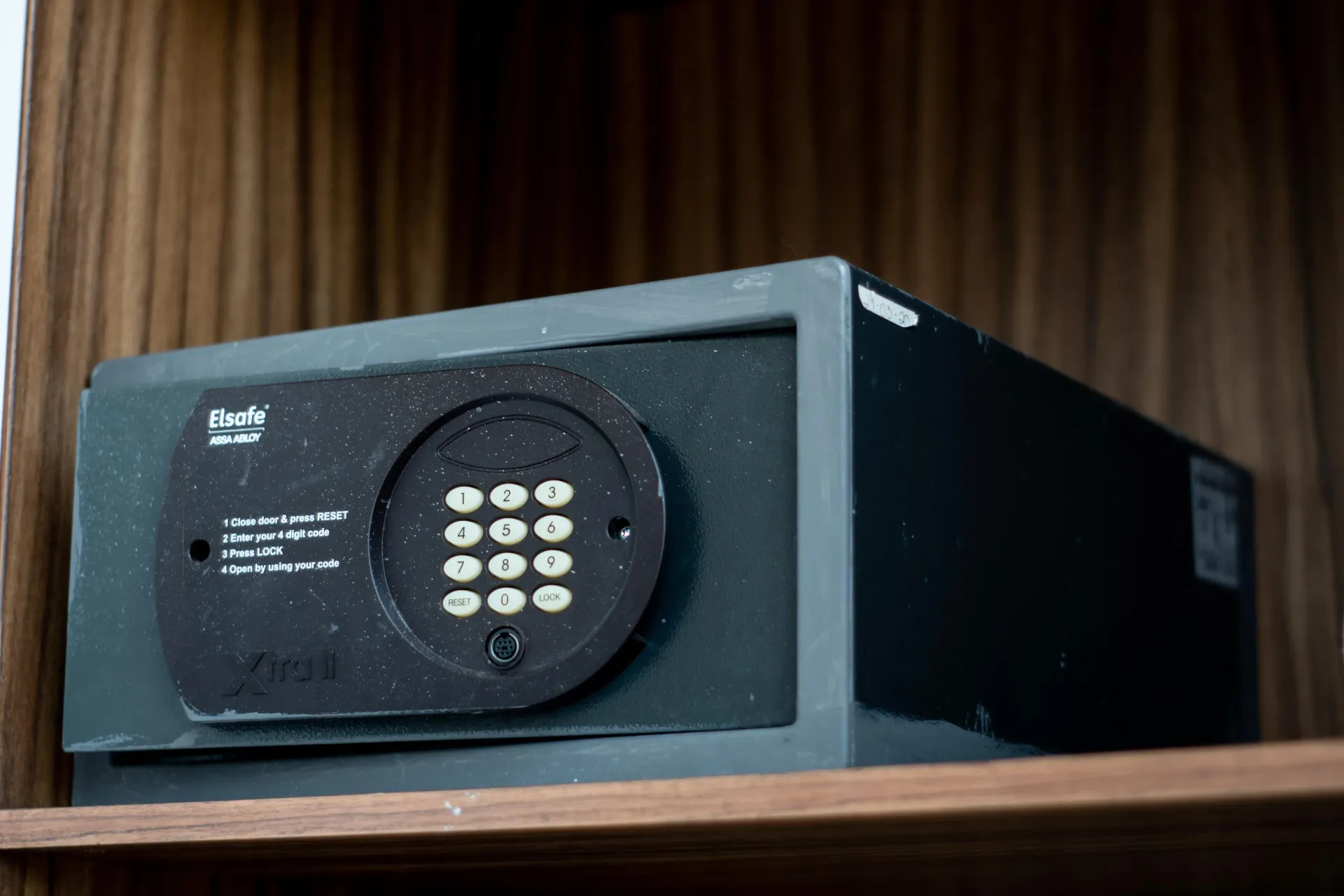 Caja fuerte electrónica negra con teclado sobre un estante de madera contra un fondo de pared a rayas.
