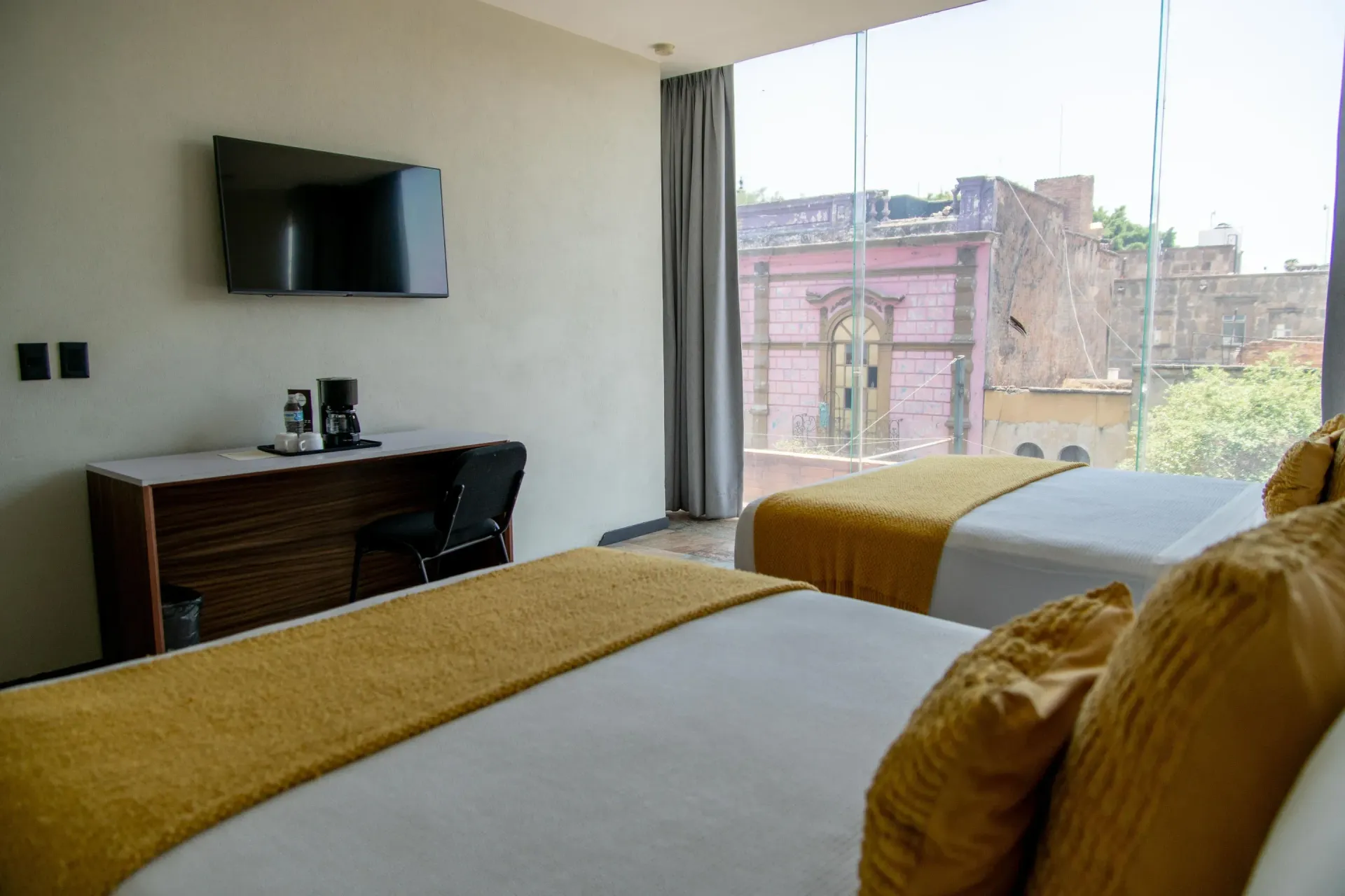 Habitación de hotel con dos camas, TV, escritorio y una gran ventana con vistas a una calle.