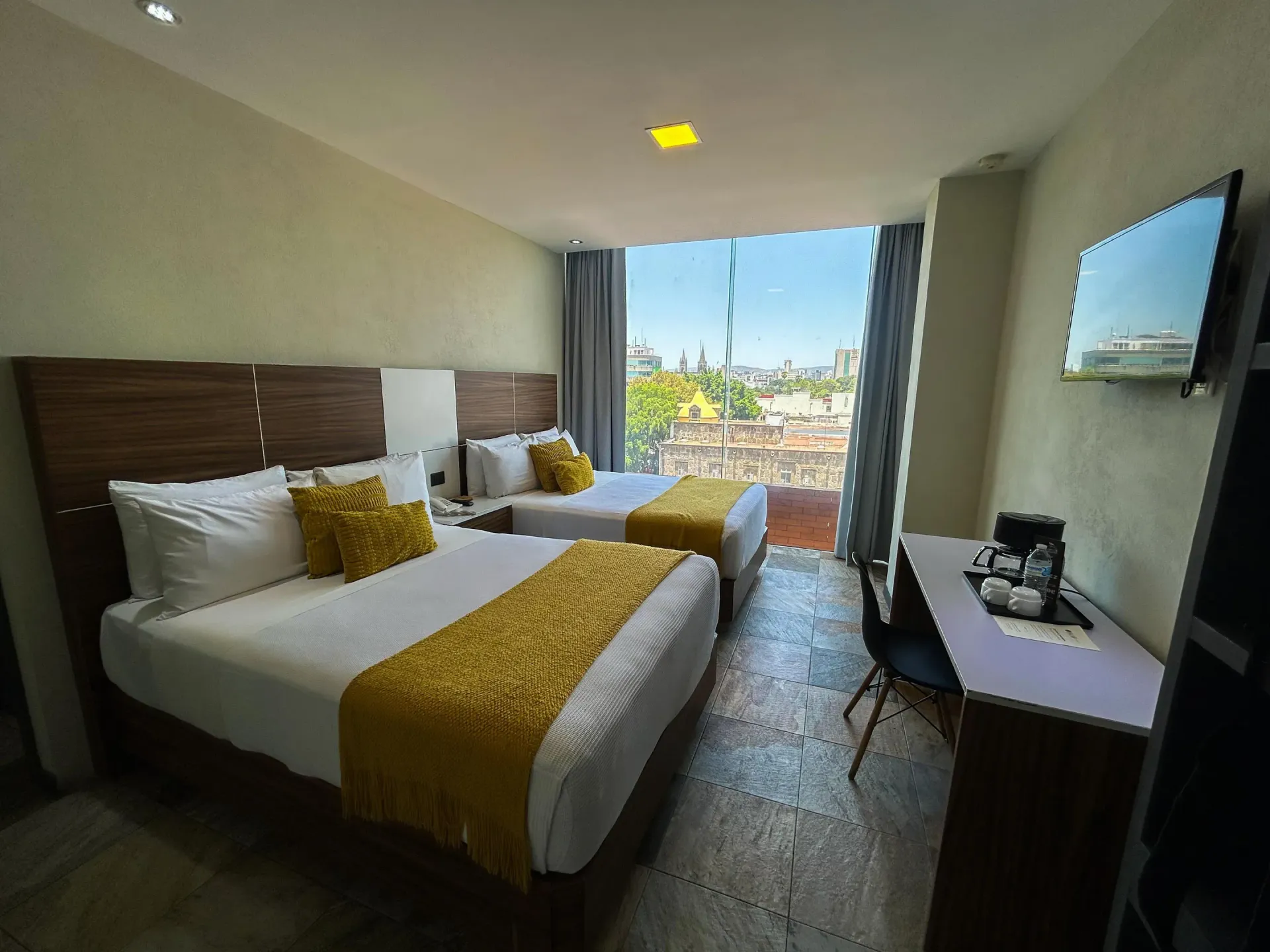 Habitación de hotel con dos camas, escritorio y balcón con vistas a la ciudad.