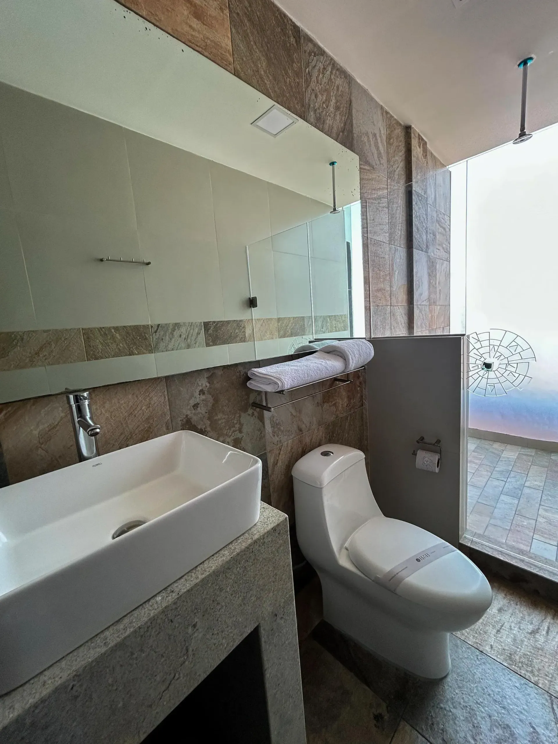 Baño moderno con lavabo de piedra, inodoro, espejo grande y zona de ducha de cristal.