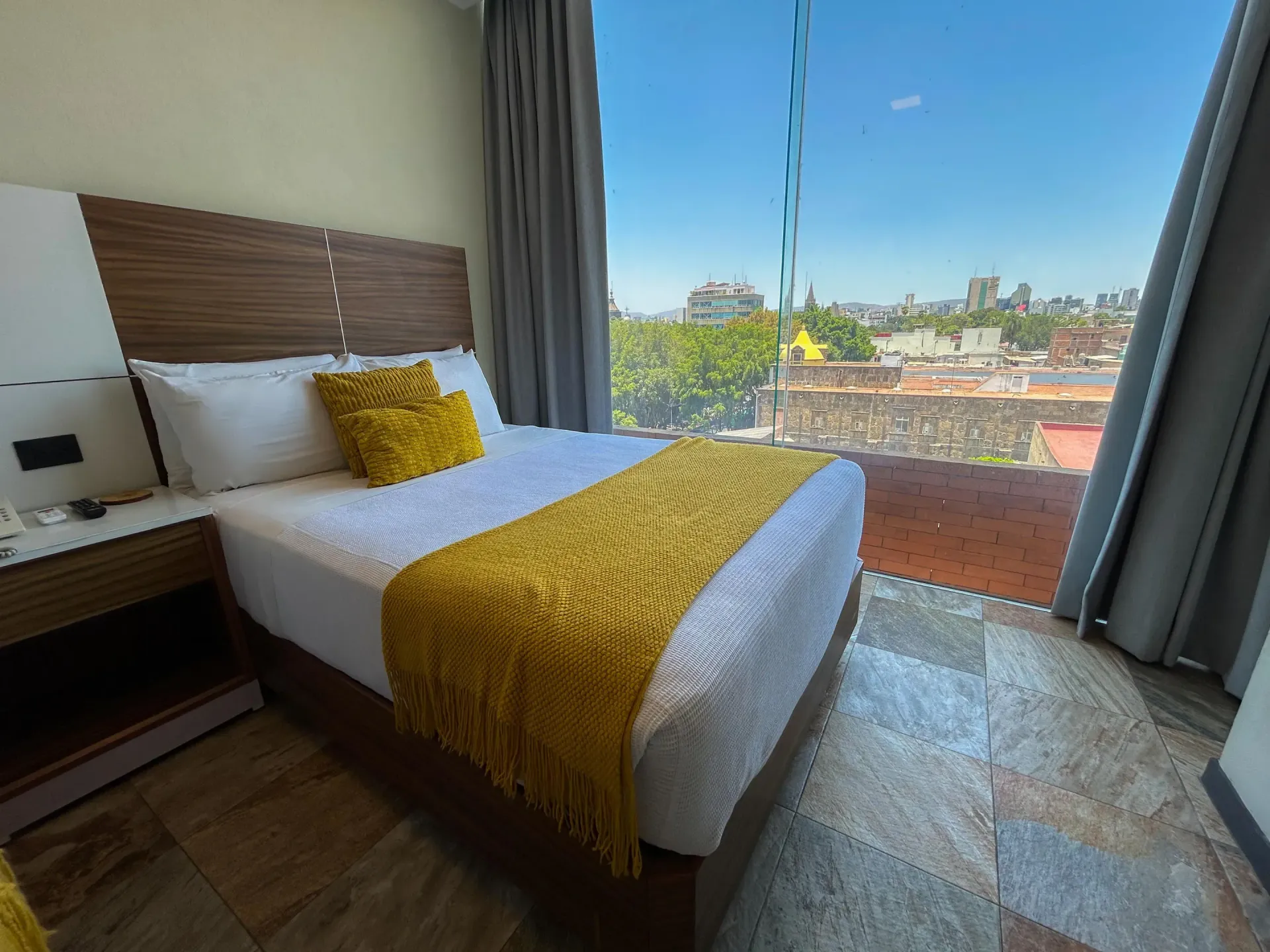 Dormitorio moderno con cama, manta amarilla, ventanal de suelo a techo y vistas a la ciudad.