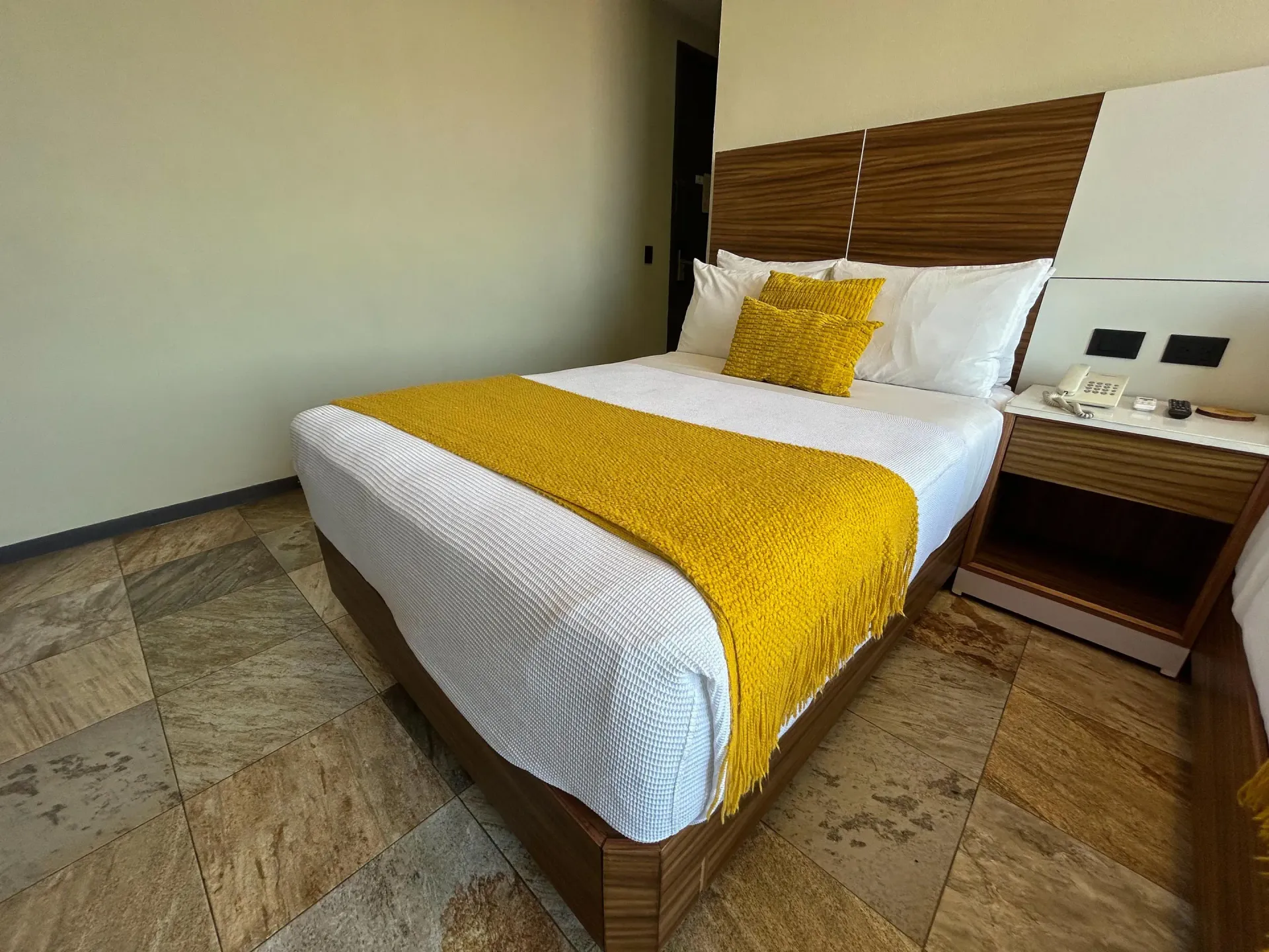 Cama bien hecha con ropa de cama amarilla en una habitación de hotel moderna con cabecero y mesita de noche de madera.