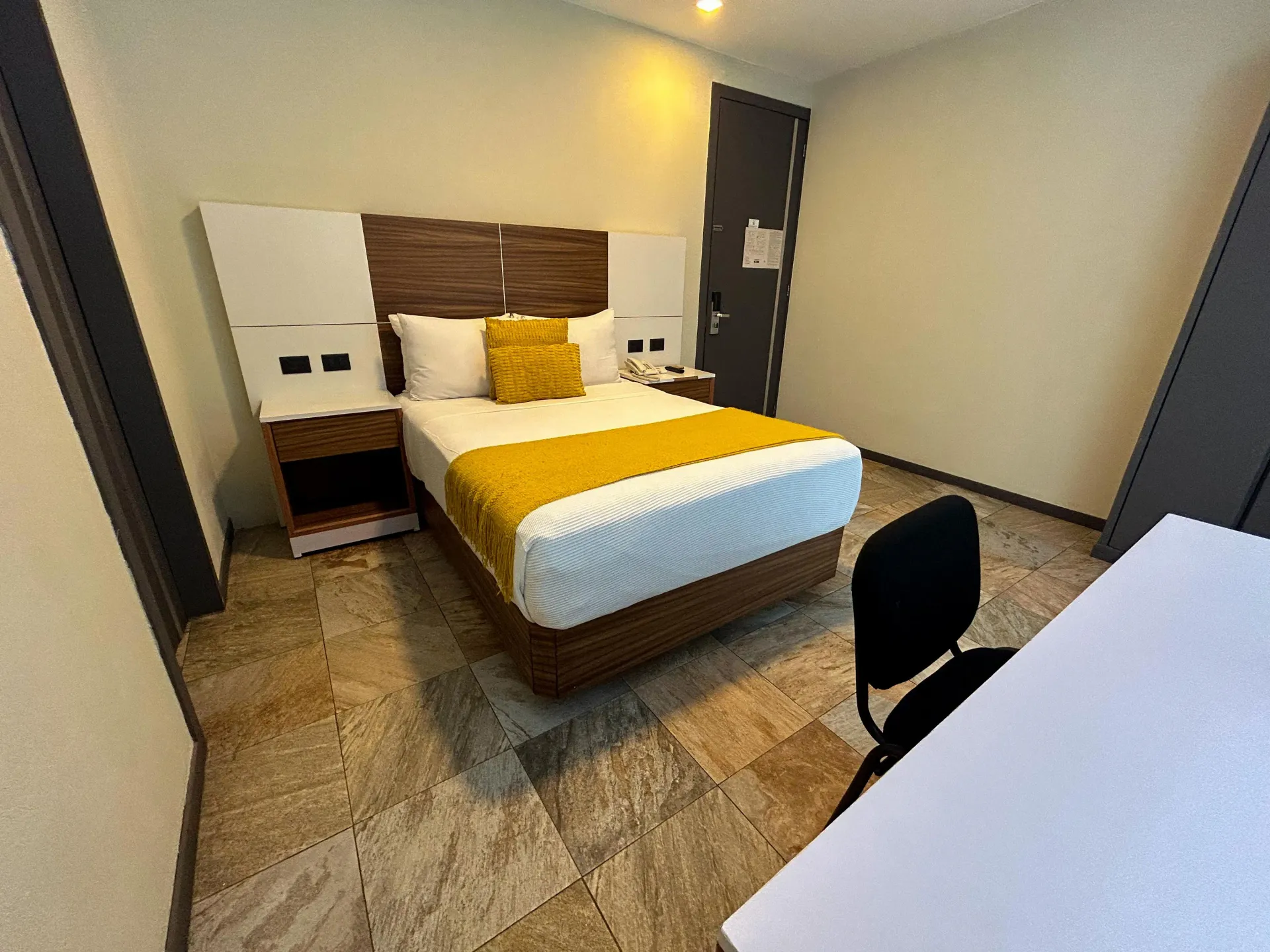 Habitación de hotel moderna con cama doble, ropa de cama amarilla, mesitas de noche y silla de escritorio.